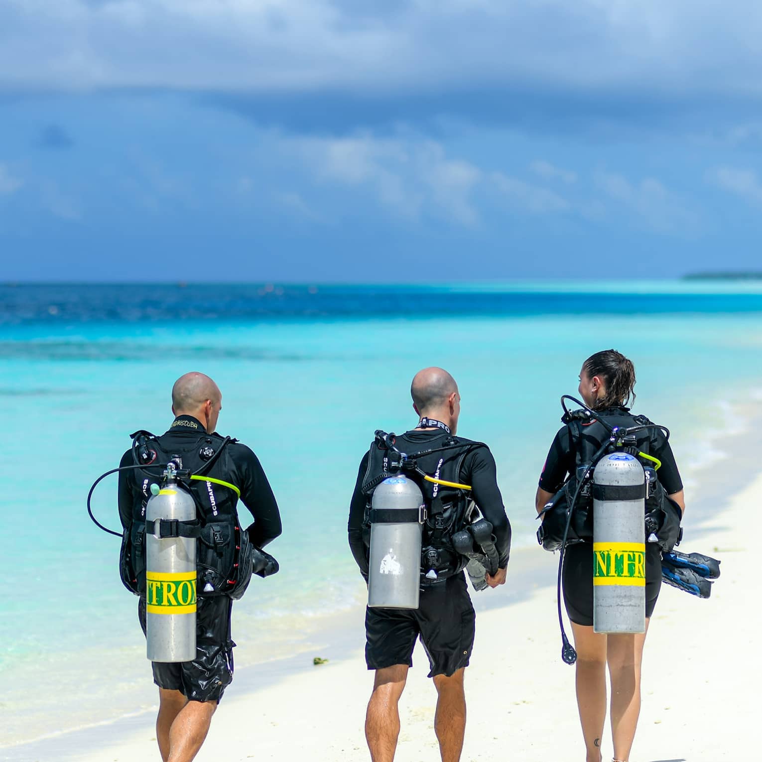 Three people wearing wetsuits and scuba tanks, walk down a white sandy beach beside the clear shimmering turquoise ocean.