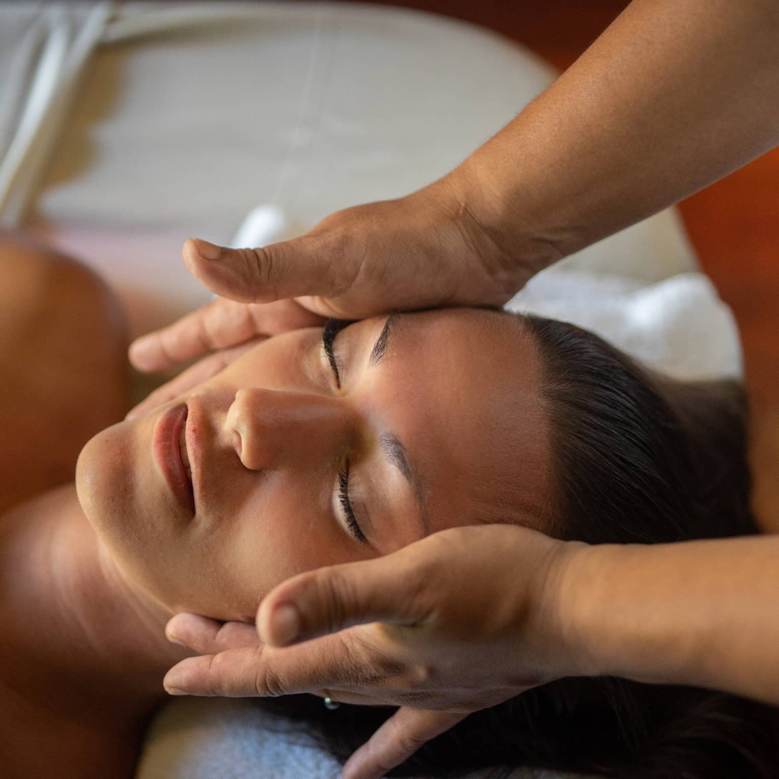 Woman lays on massage table with eyes closed as hands massage her cheeks and chin