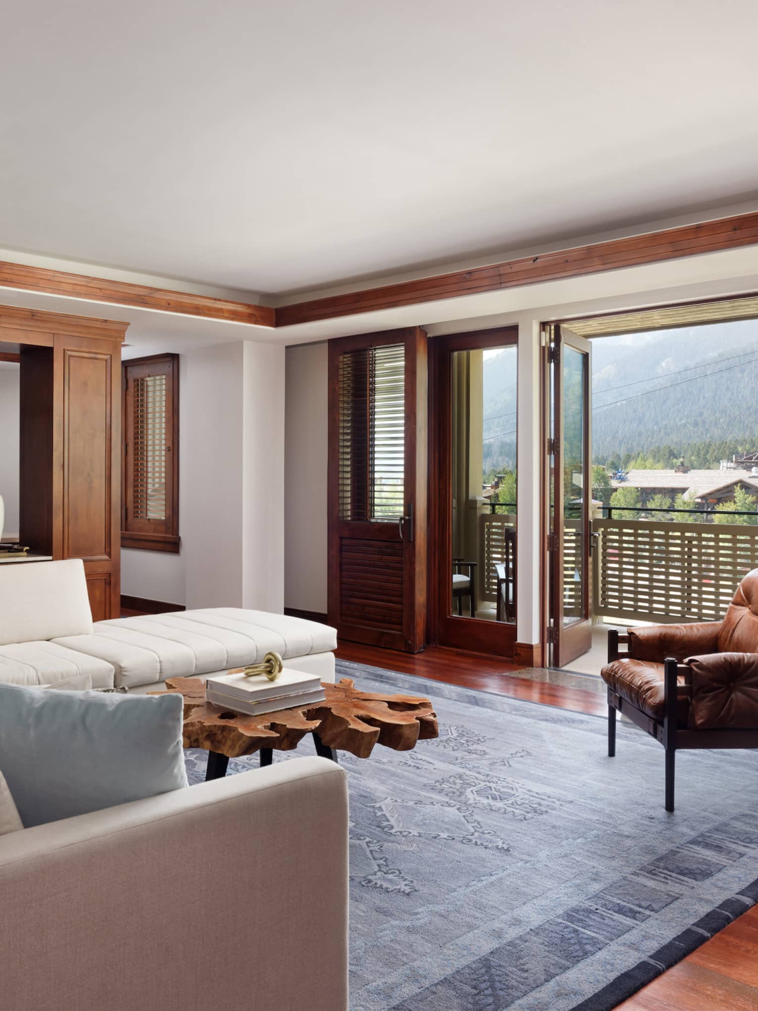 Suite living room with a cream sectional sofa, rustic wood coffee table, leather armchair and large doors opening to a balcony with a mountain view