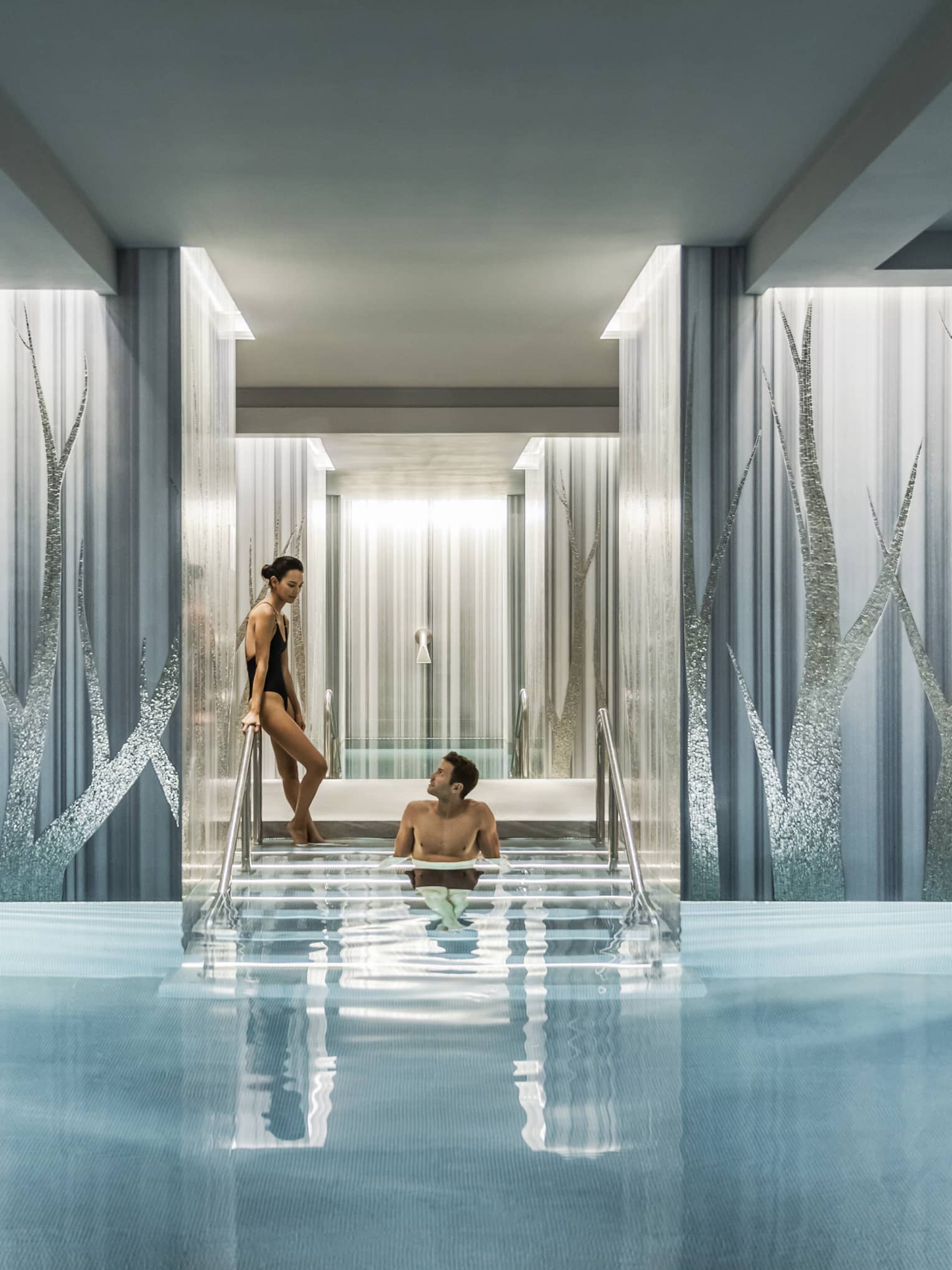 Woman in black swimsuit enters serenity pool while man sits on steps between blue and silver columns