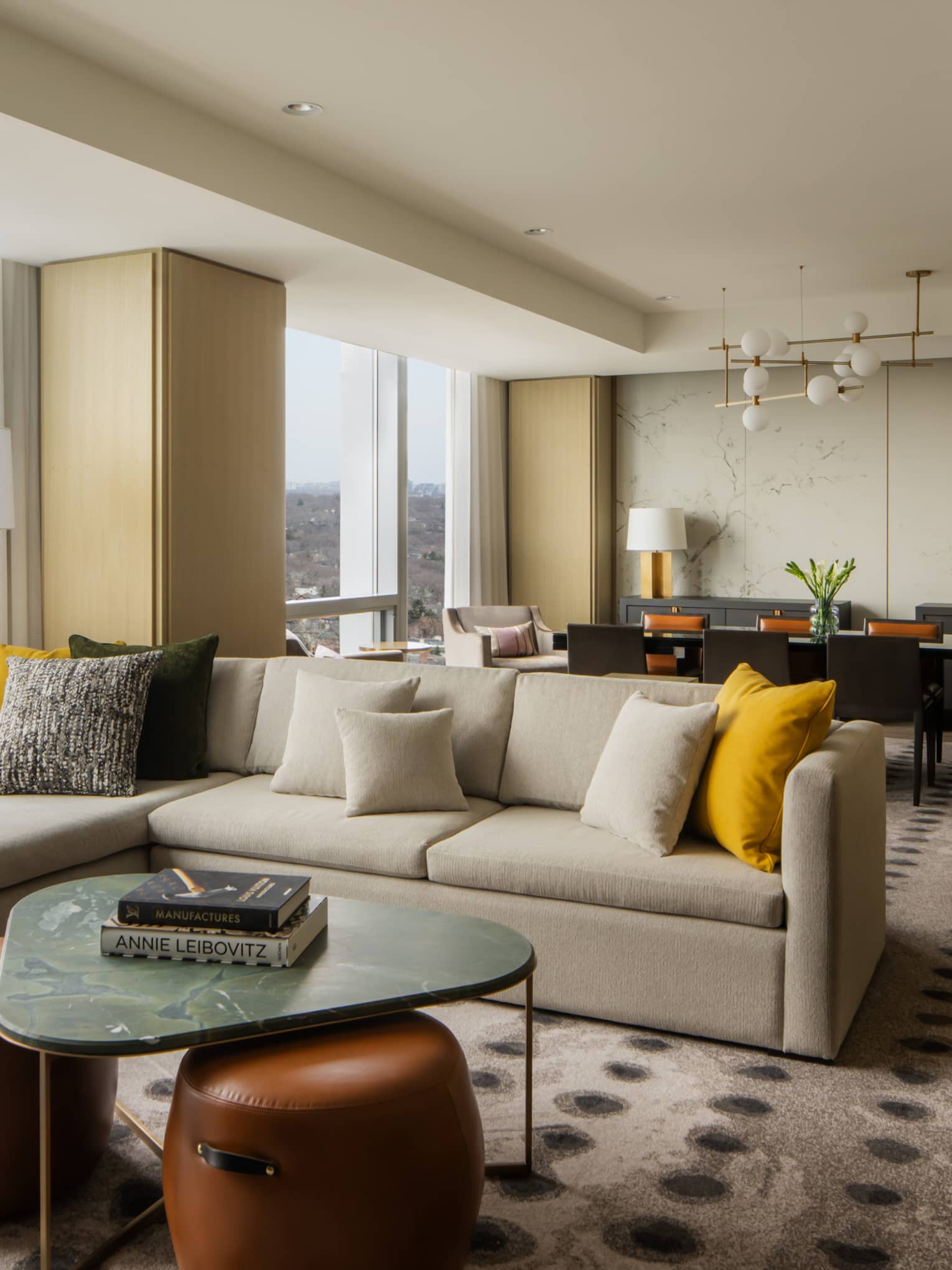 Spacious suite living area with a large sectional sofa, yellow and gray pillows, marble coffee table, and a dining area with elegant décor and city views