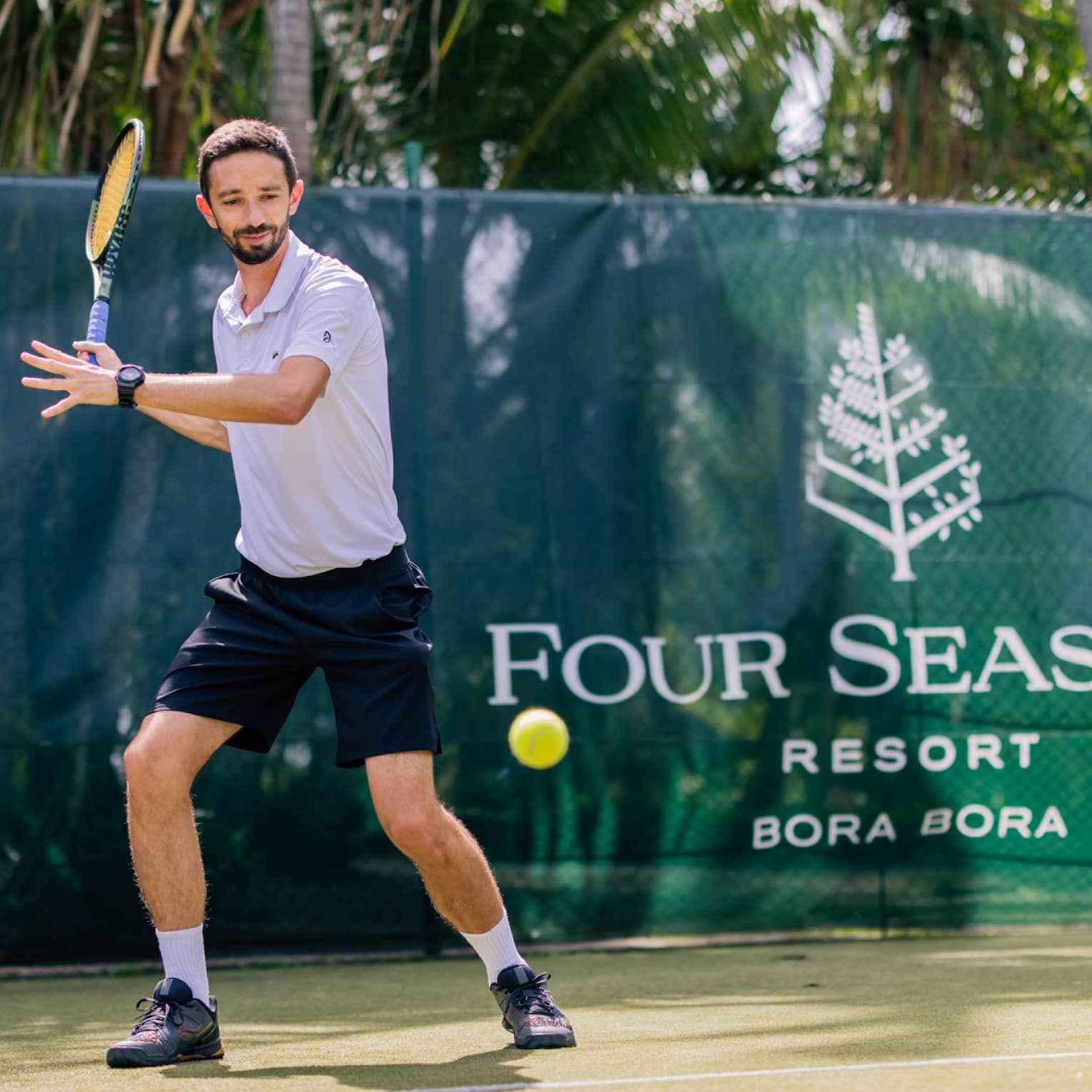 Standing behind the baseline on a court surrounded by palm trees, a tennis player prepares to hit a forehand.