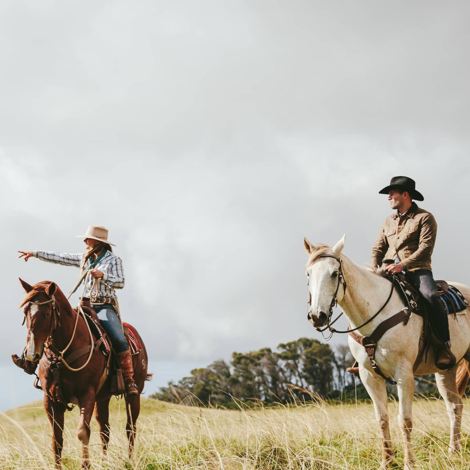 A couple riding horses, looking out at the long grass fields, with grassy hills and trees in the distance on a cloudy day.