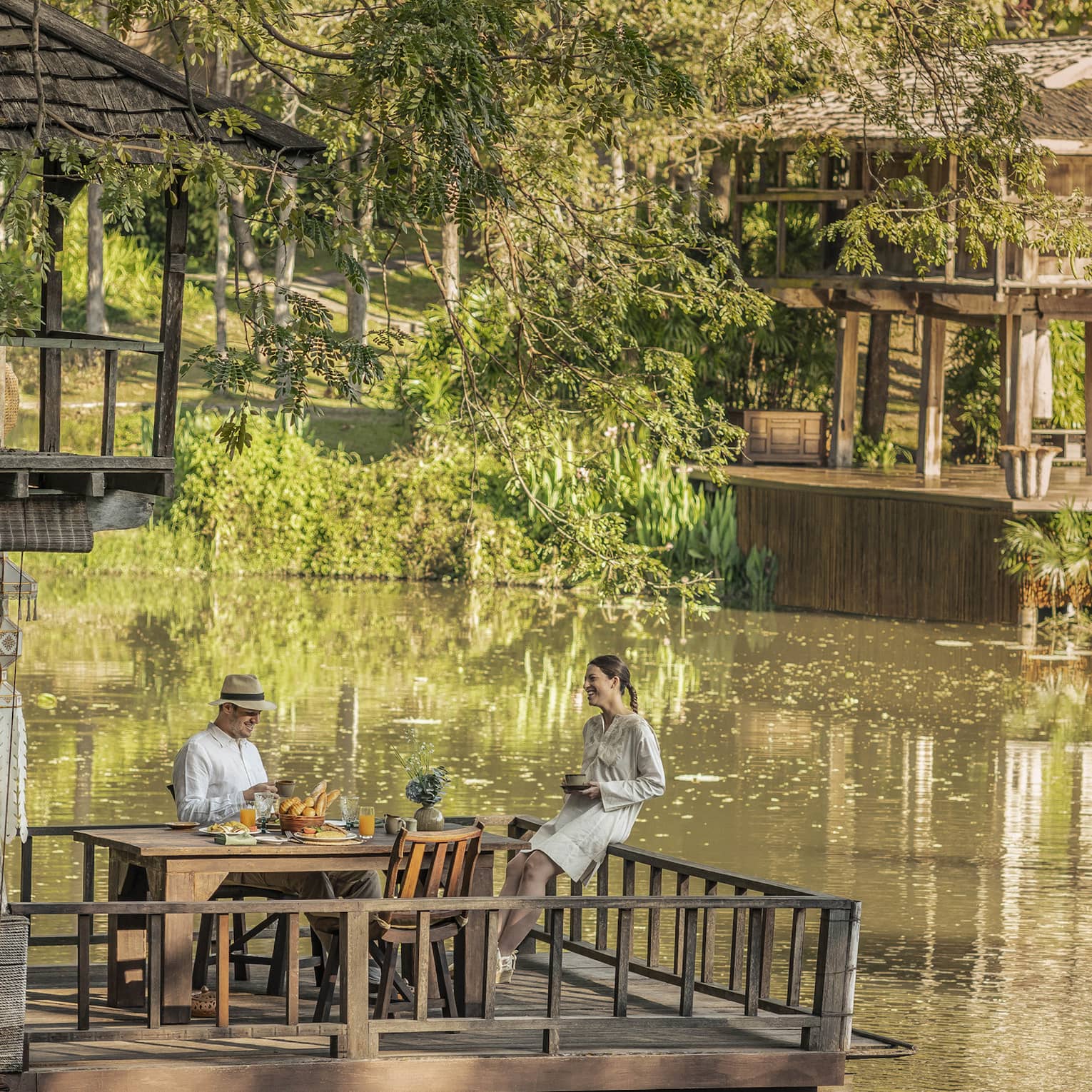 Two guests at a rustic table, on a platform in one of two wood huts on stilts perched atop a calm river bordered by greenery.
