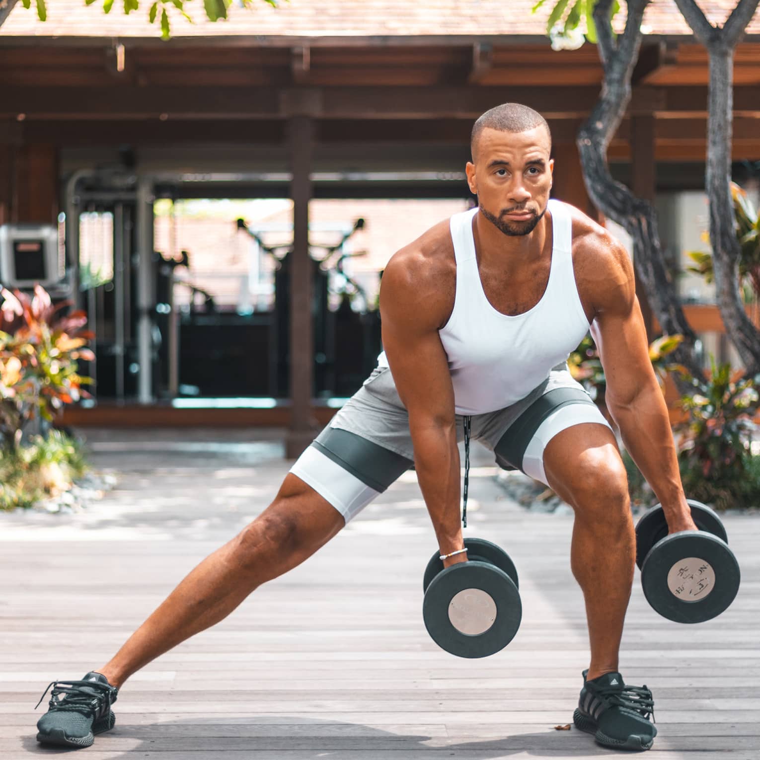 Man in white tank top and shorts doing side lunge on outdoor decking, holding weights