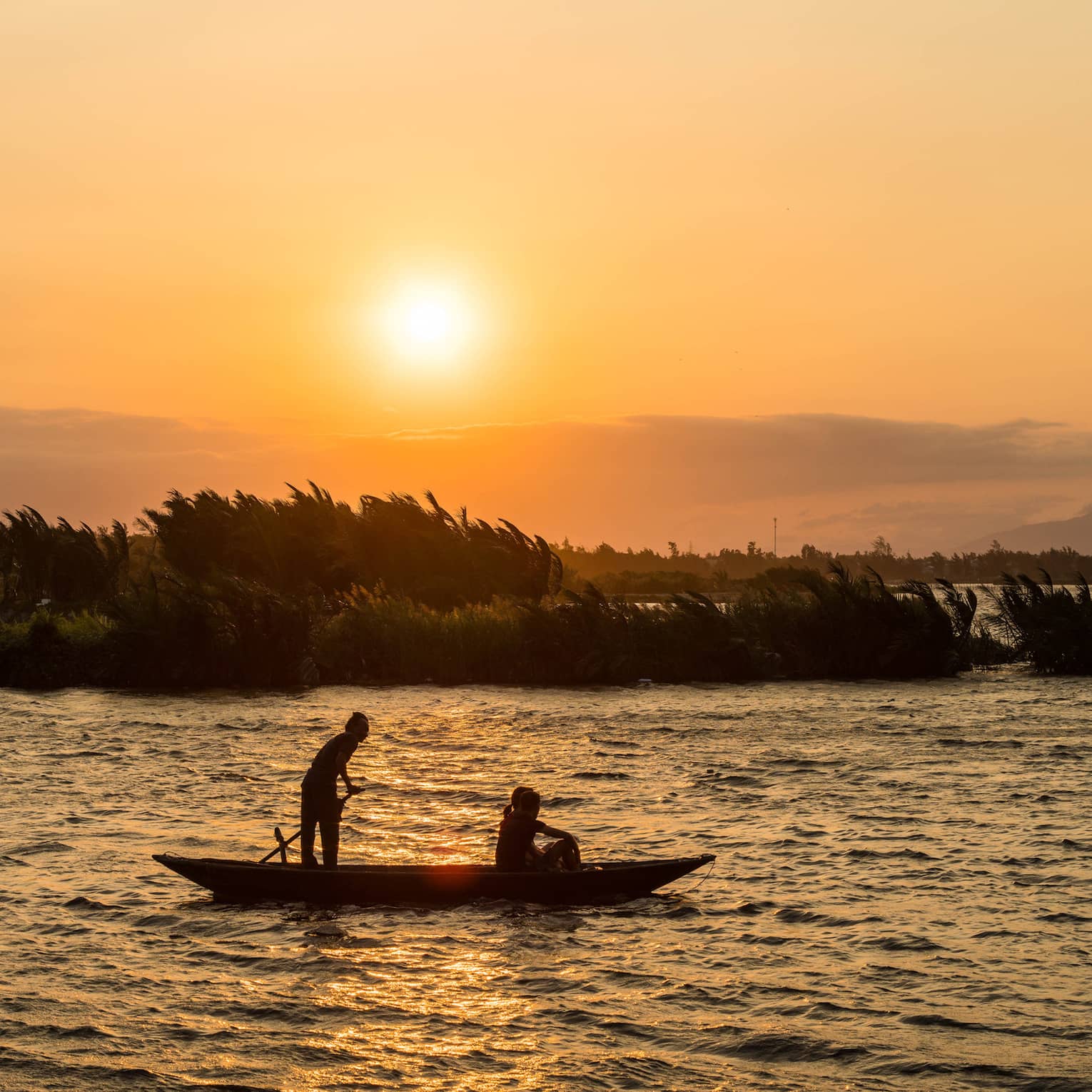 Silhouetted against a hazy orange sunset and windblown trees, two people in a bamboo basket boat drift on rippling water.