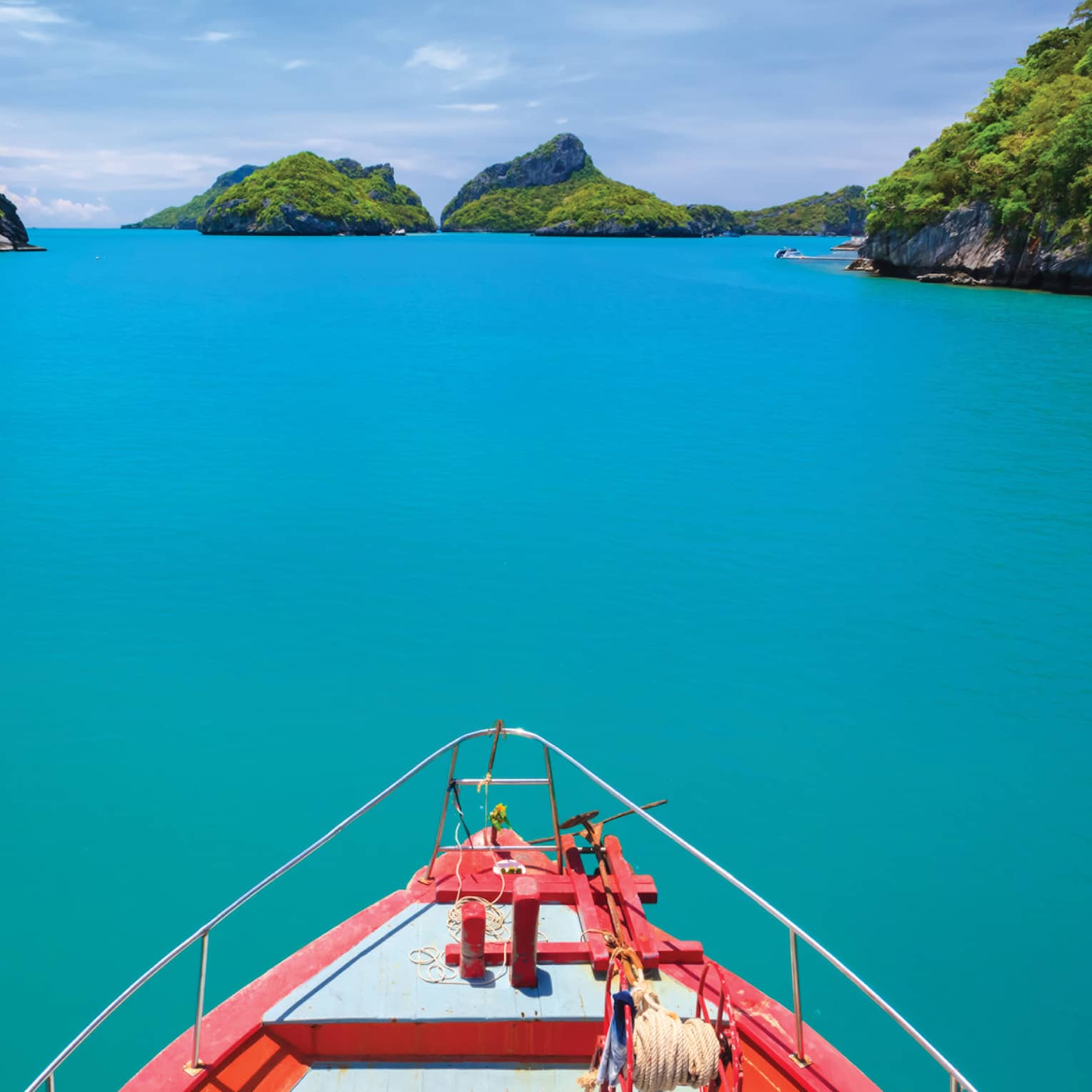 A view from the bow of a wooden boat overlooking calm, turquoise waters and several rocky tree-lined islands all around.
