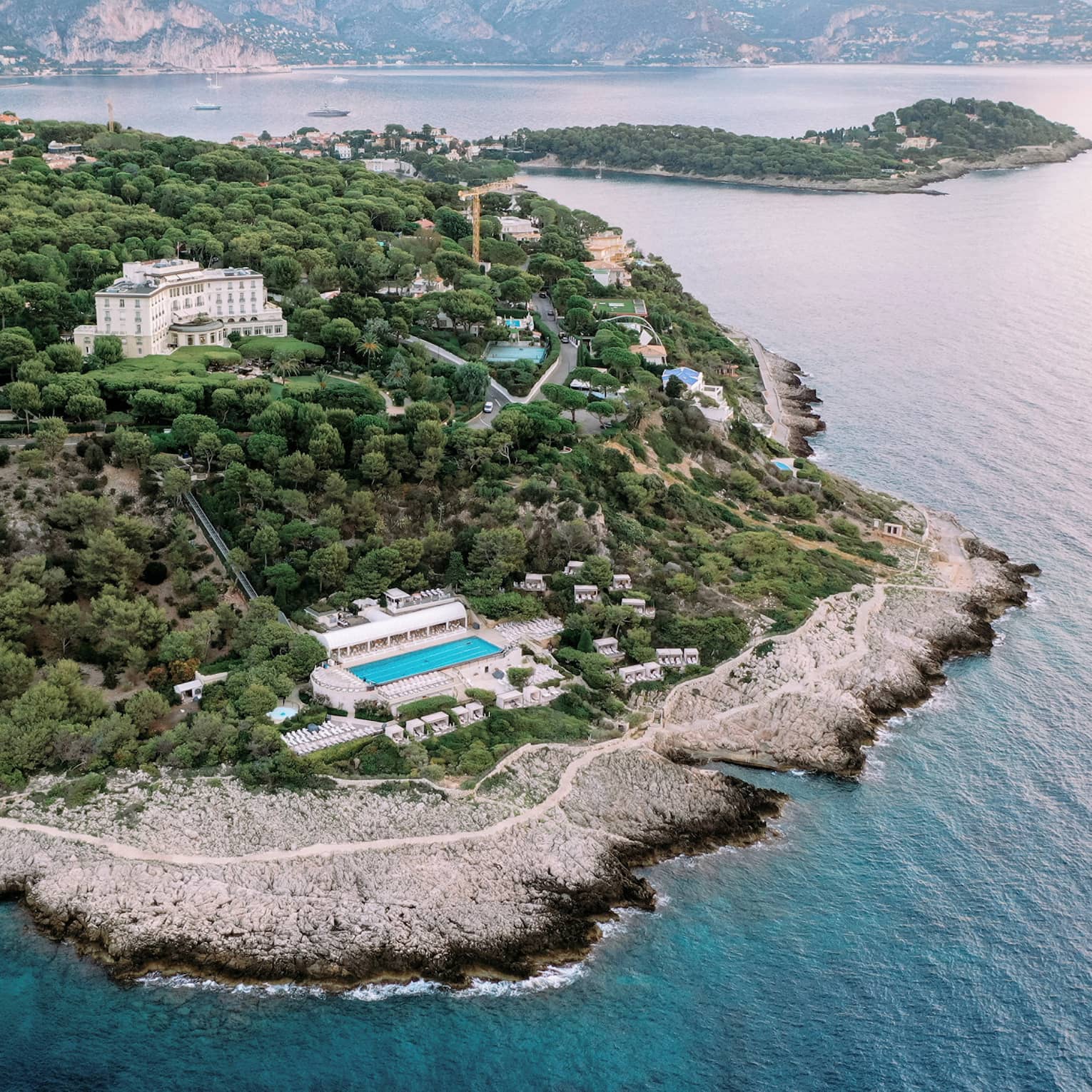 Aerial view of buildings and pools nestled among verdant trees on a sloping hillside along an extensive, rugged coastline.
