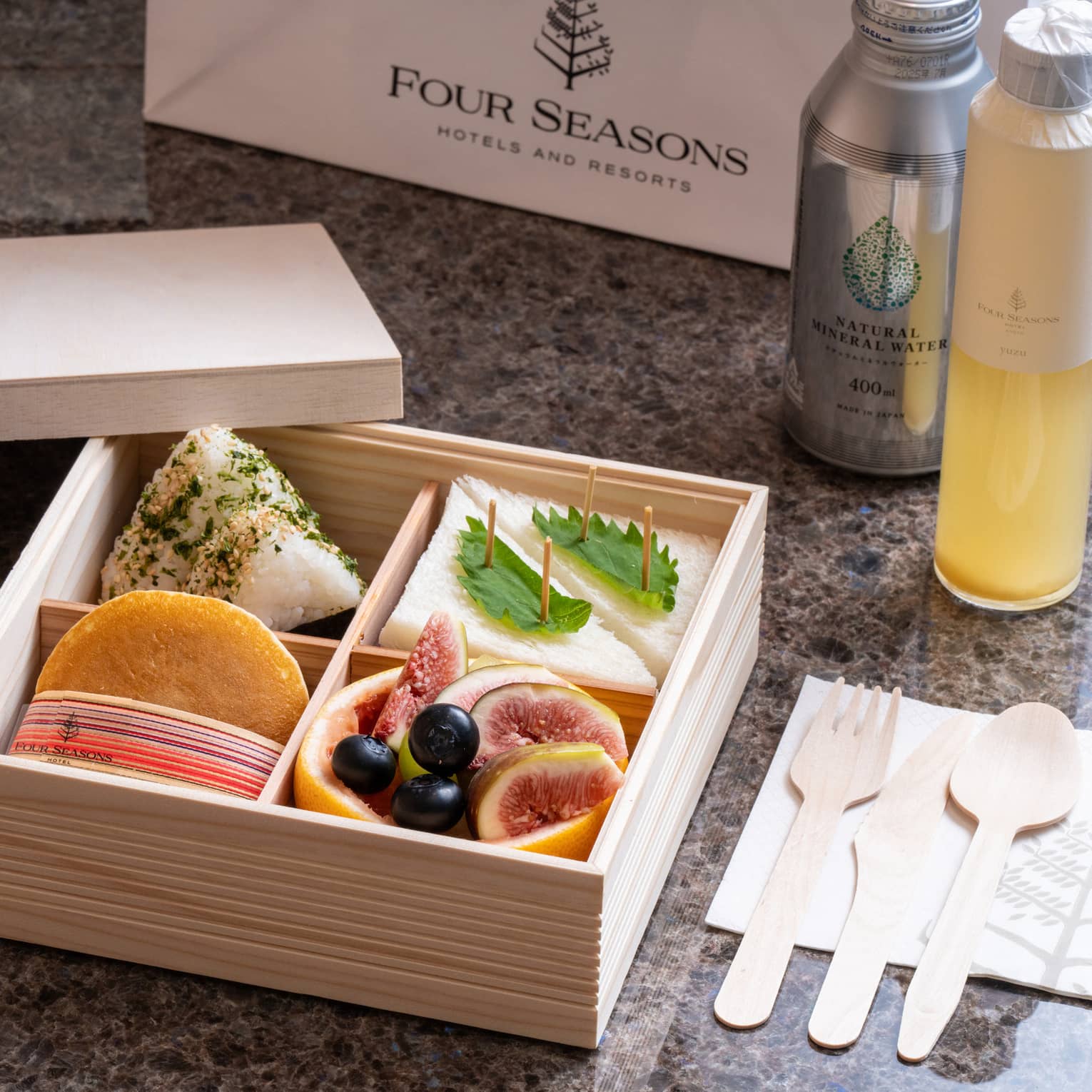 A wooden Four Seasons bento box with four compartments holding sandwiches, fruits and delicacies, beverage bottles nearby.