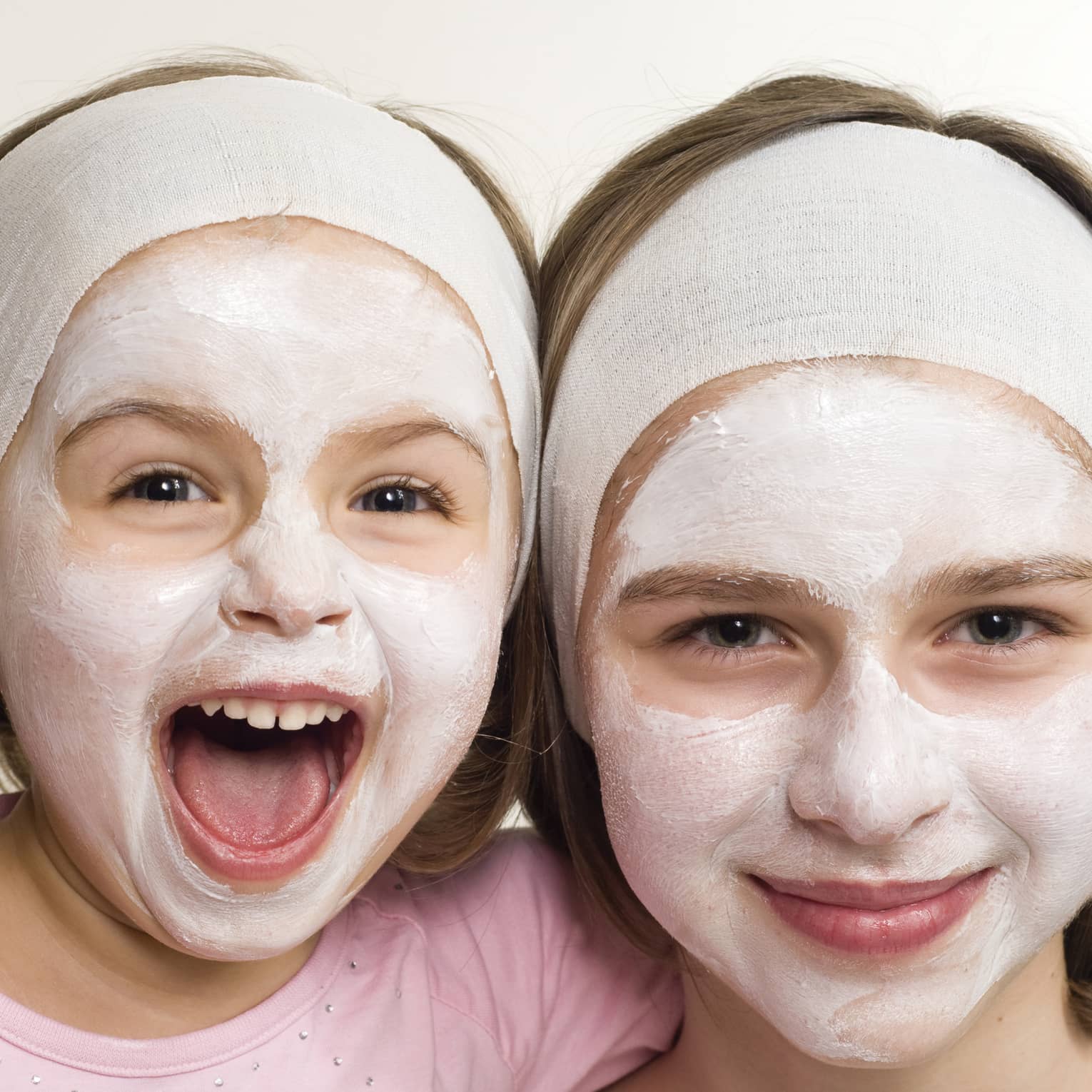 Two smiling children wearing headbands and facial masks, one laughing joyfully, showcasing a fun skincare moment.