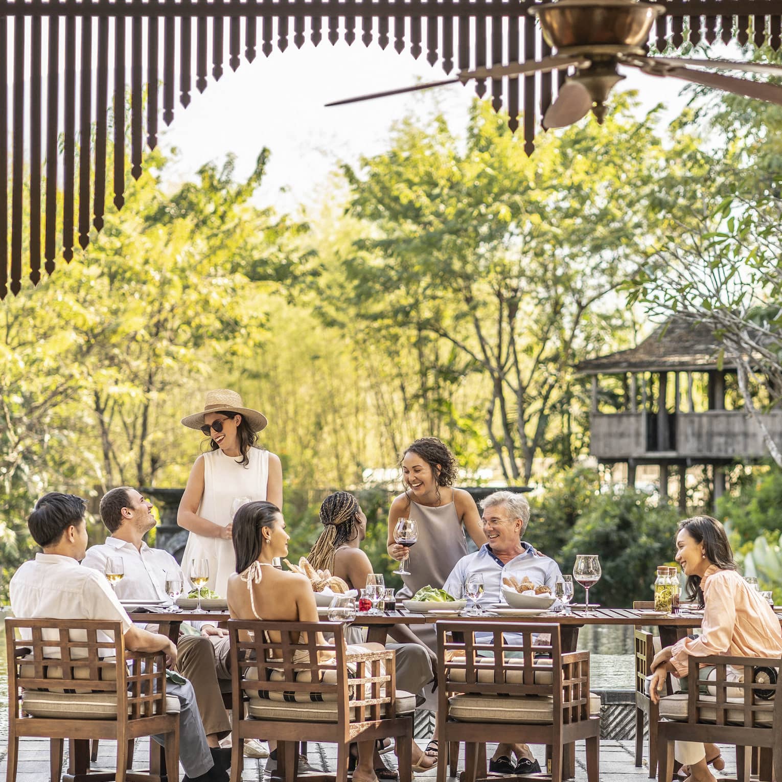 A smiling group around a rustic patio set with wine and appetizers, as a chef barbecues corn and light filters through trees.