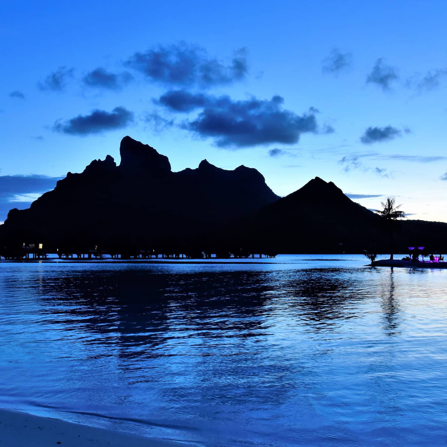 A row of overwater bungalows and a rocky mountain are silhoutted against the blue, twilit sky and above the smooth ocean.