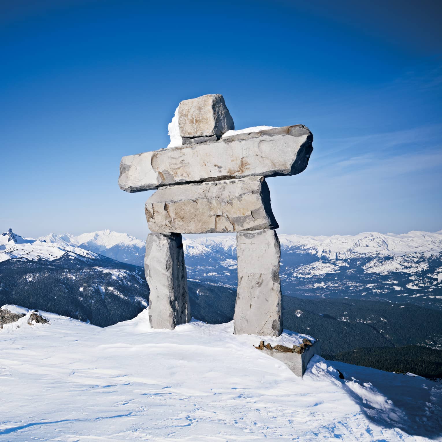 Inukshuk stone sculpture towering at top of mountain against blue sky backdrop, snow-capped mountains