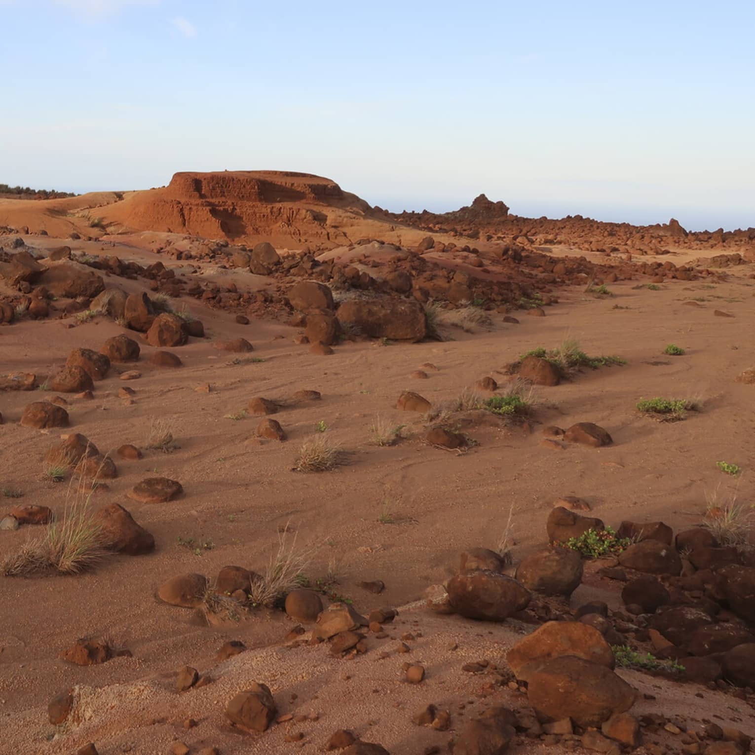 A desert landscape of red boulders, rocks, sand and small rocky hills with the occasional green vegetation growing.