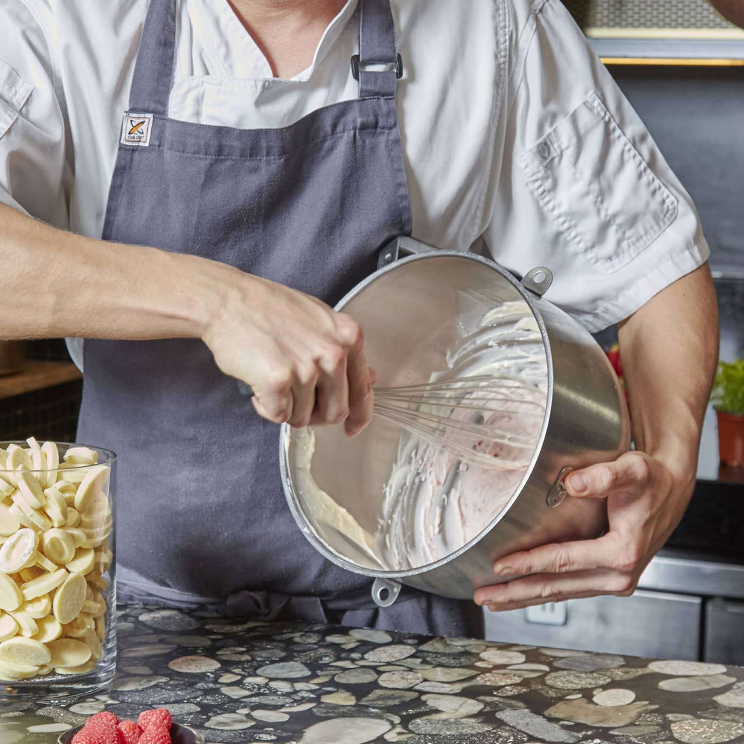 An aproned chef whisks cake batter in a bowl over a pebbled countertop, by a rolling pin and jar of white chocolate pieces.