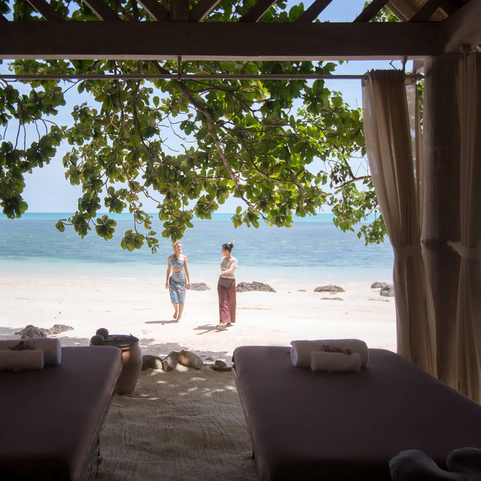 Couples massage beds under cabana in front of two women walking on white sand beach