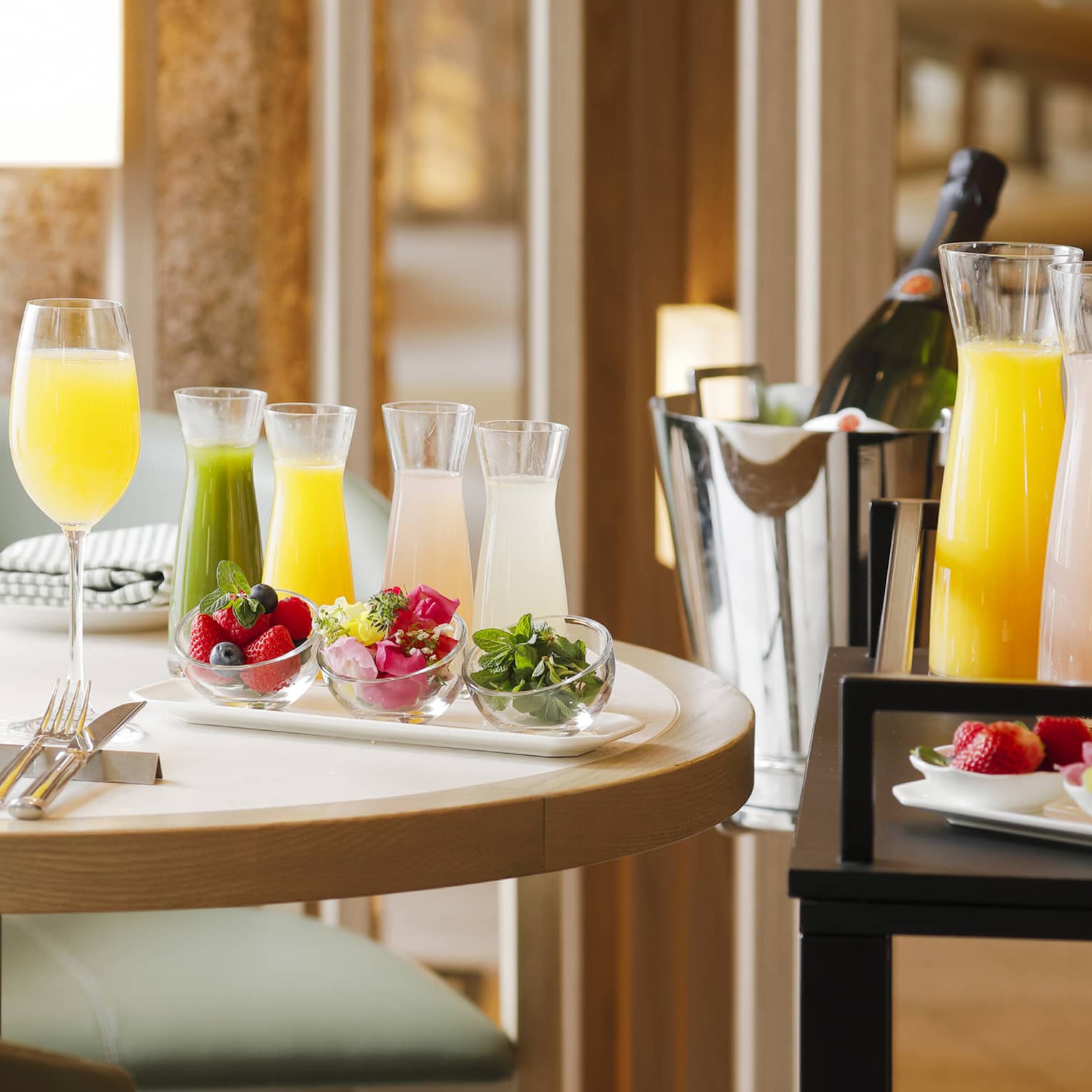 Two mimosas in long-stemmed flutes aside sets of small and large carafes of juice and bowls of fresh fruit and garnishes.