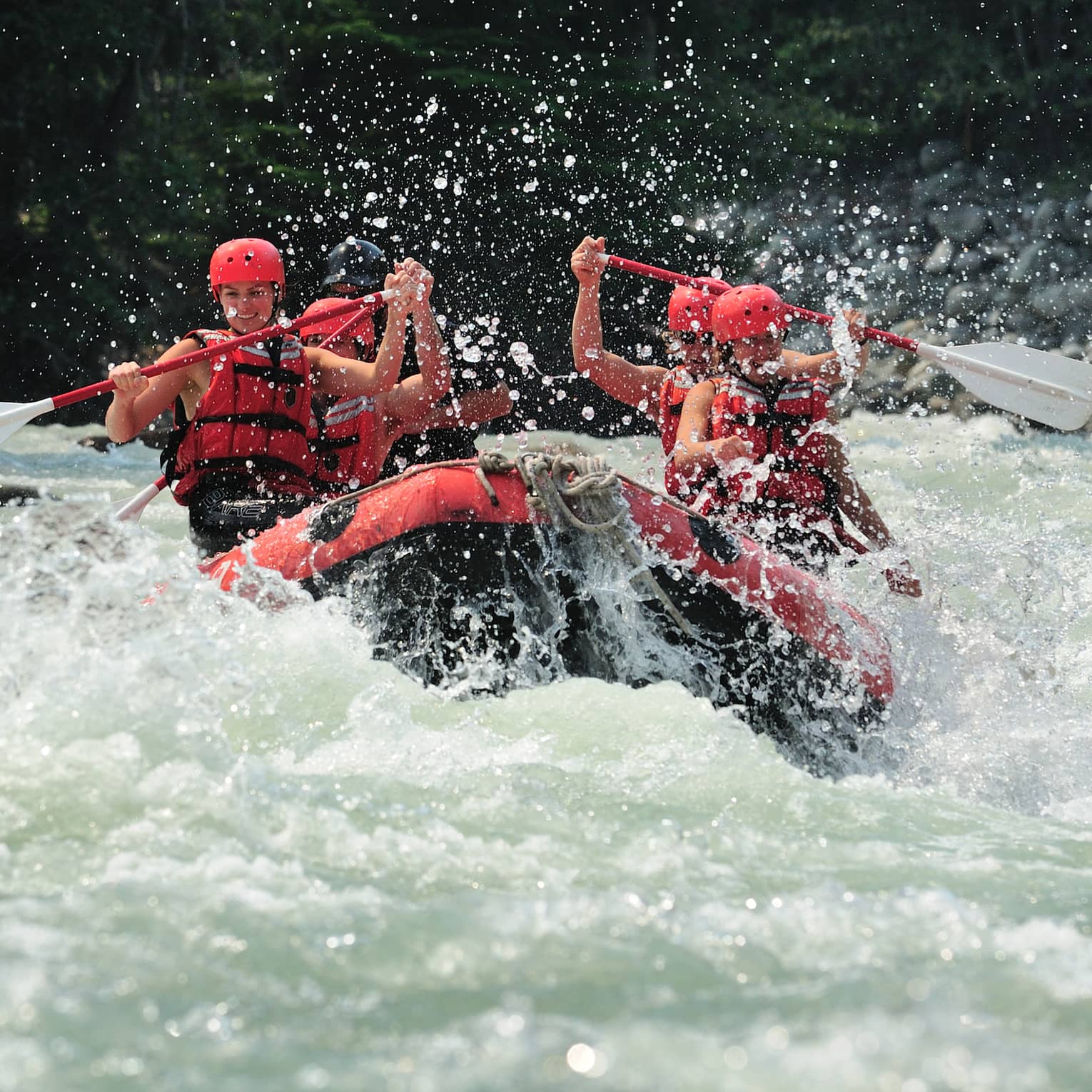 Smiling paddlers in red helmets and lifejackets steer an inflated raft through raging white water, passing evergreen trees.