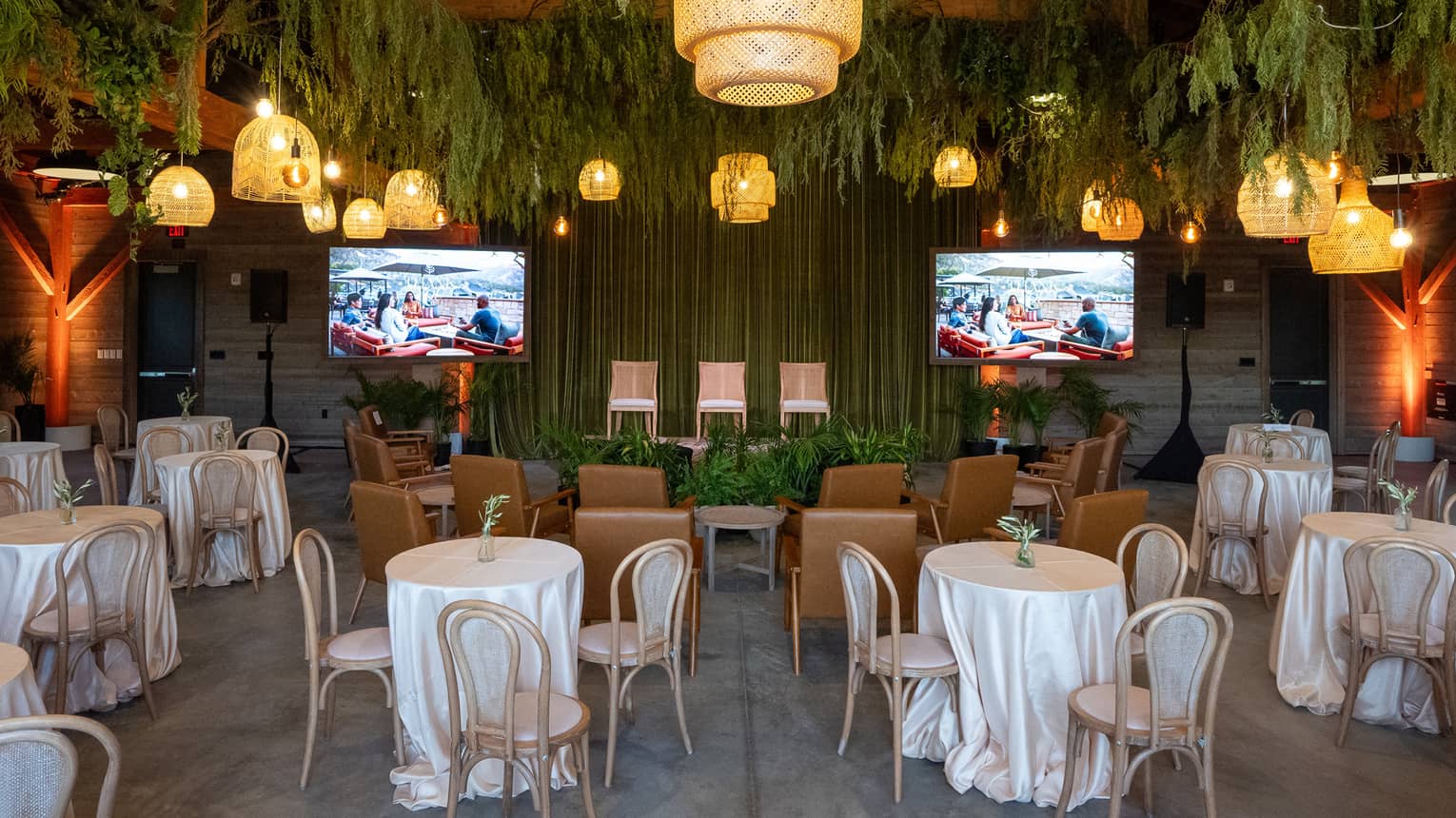 A venue with two television screens on the wall, plants hanging from the ceiling and tables with three seats