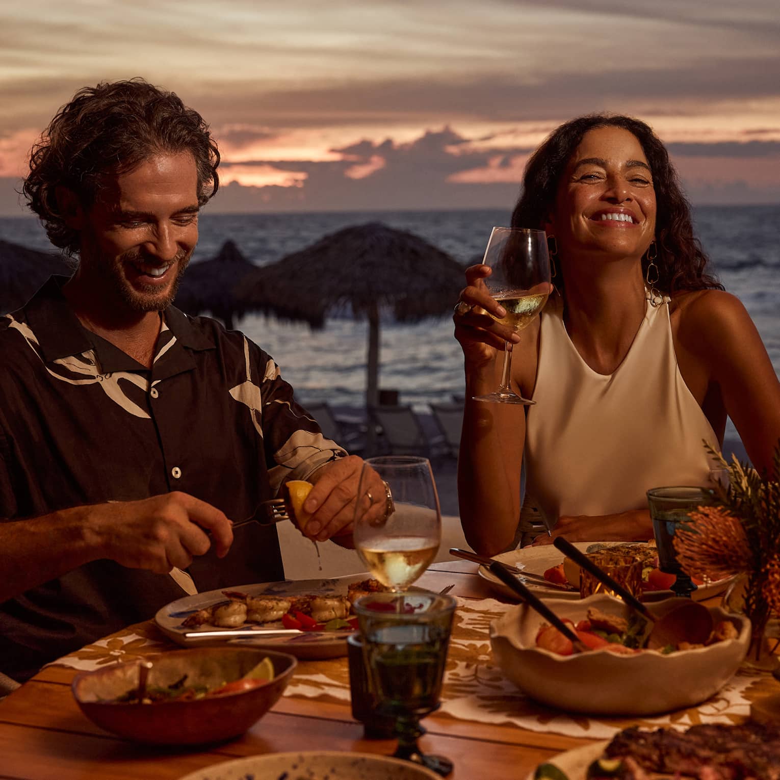 Man and woman laugh while dining at outdoor table filled with food and wine, next to beach and ocean