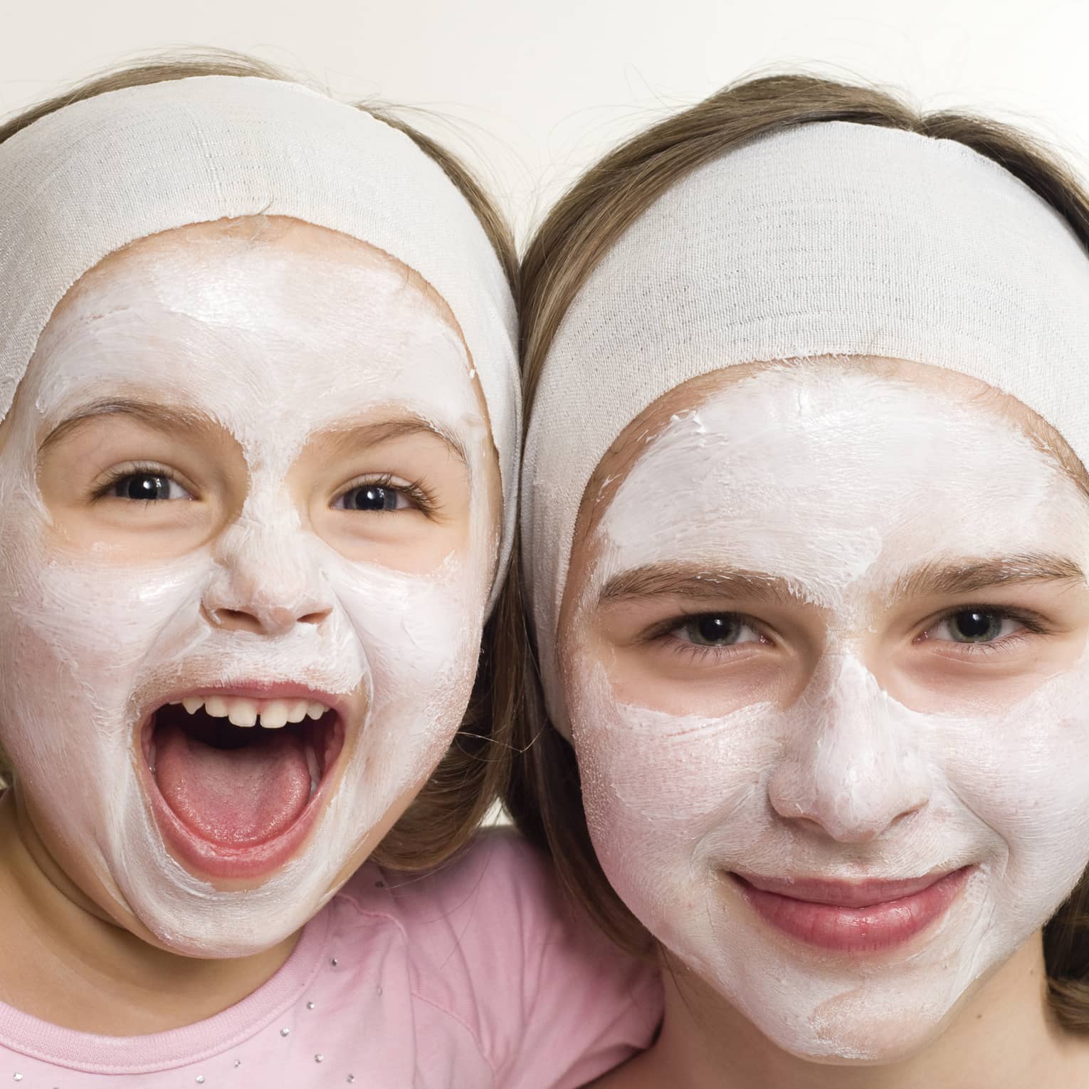 Two smiling children wearing headbands and facial masks, one laughing joyfully, showcasing a fun skincare moment.