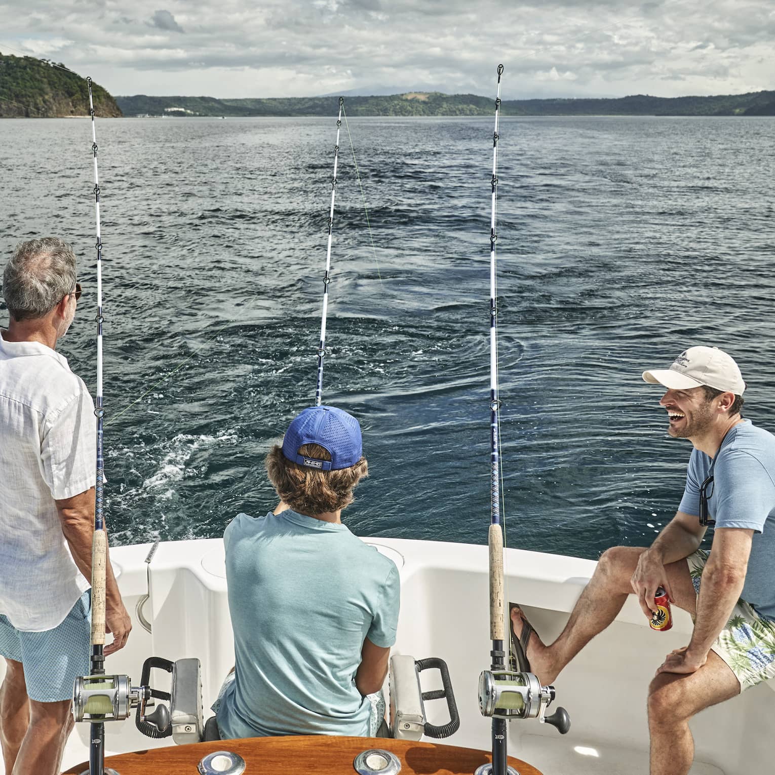 Three men of varying ages sit looking out at the water from the back of a boat outfitted with fishing rods