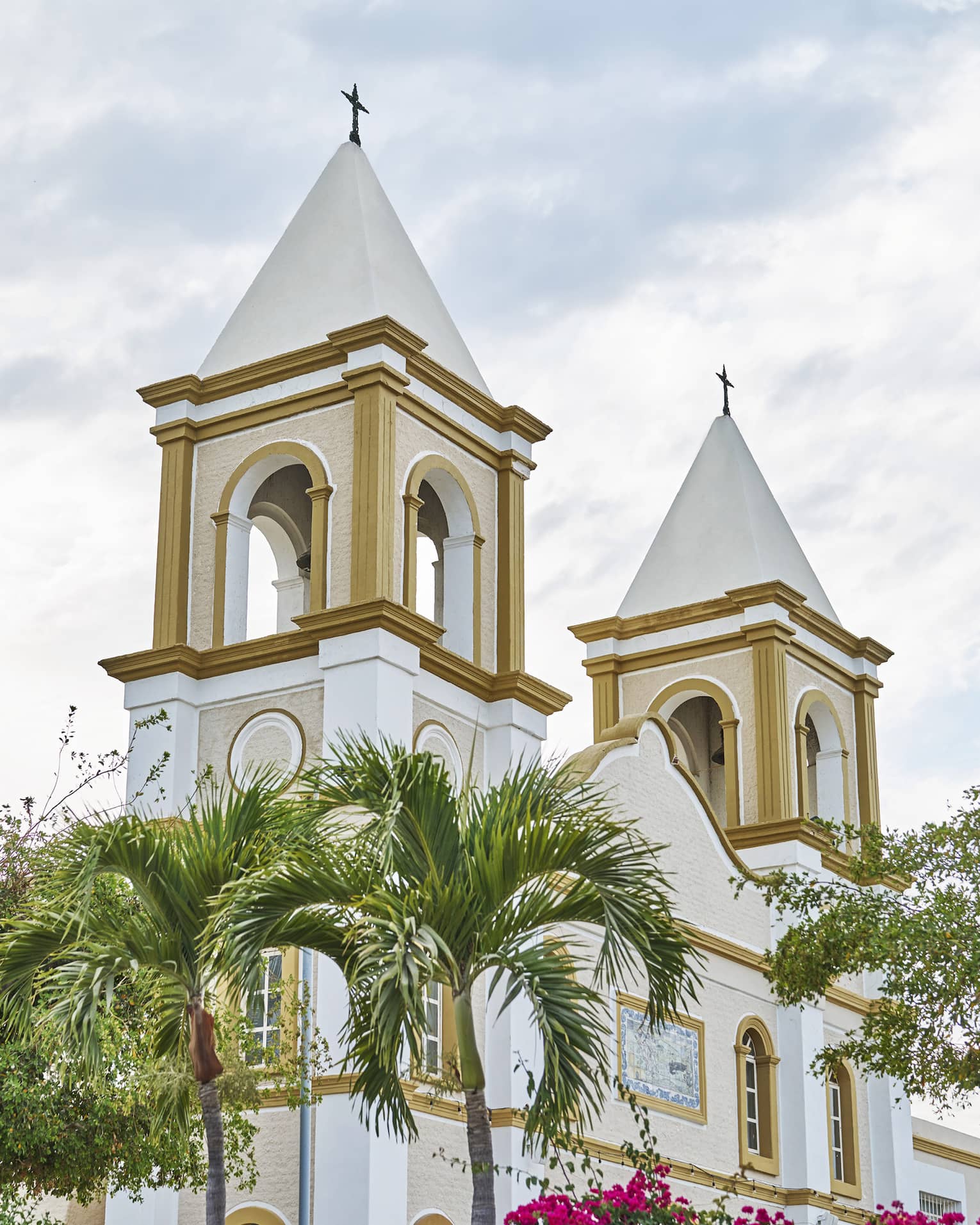 Two church bell towers rise behind palm trees and in front of a cloudy sky. They feature arched windows and gold paint.