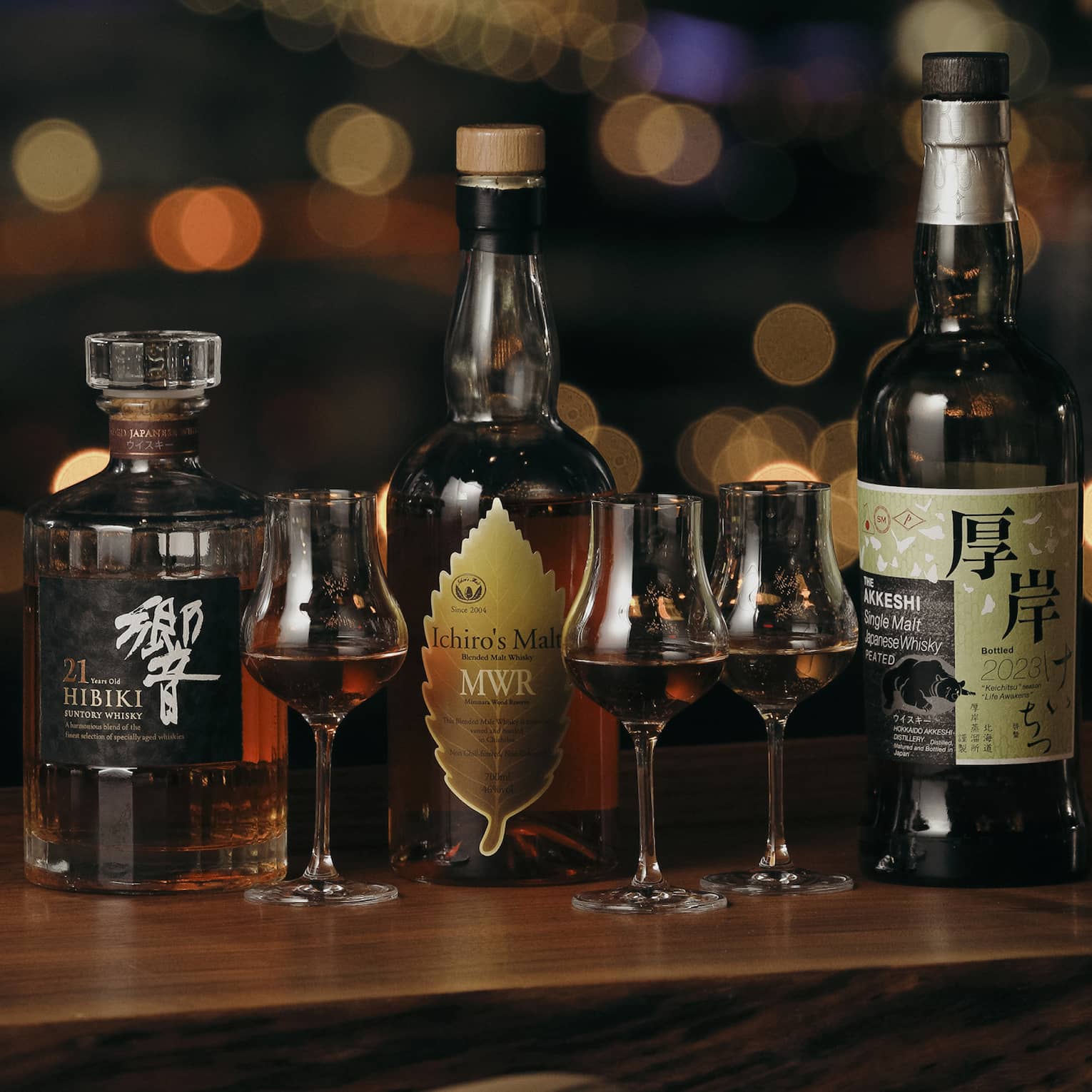Three Japanese whisky bottles with three stemmed glasses of whisky against a dark background with blurred dots of colour.