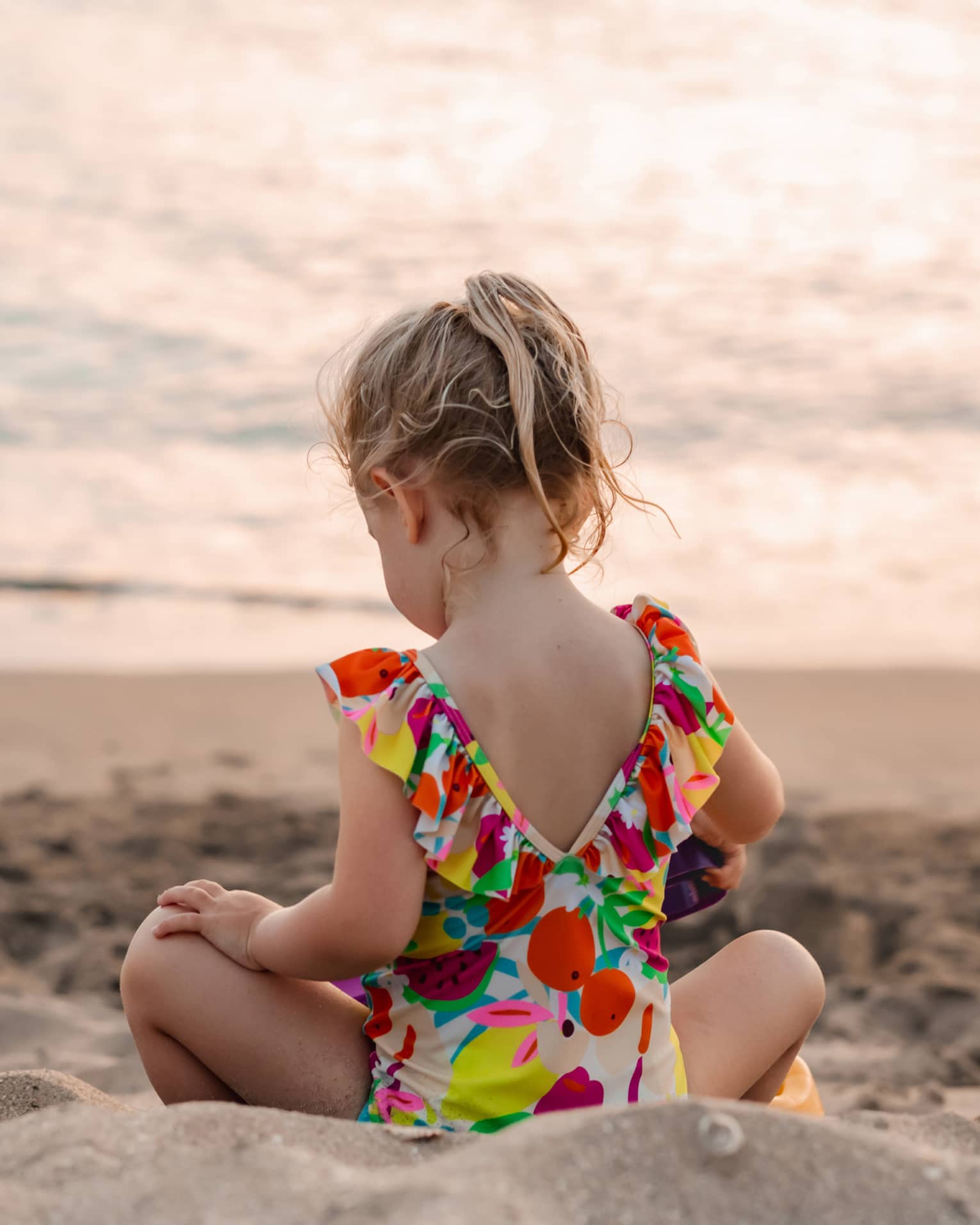 Young child sits on a sandy beach facing the water