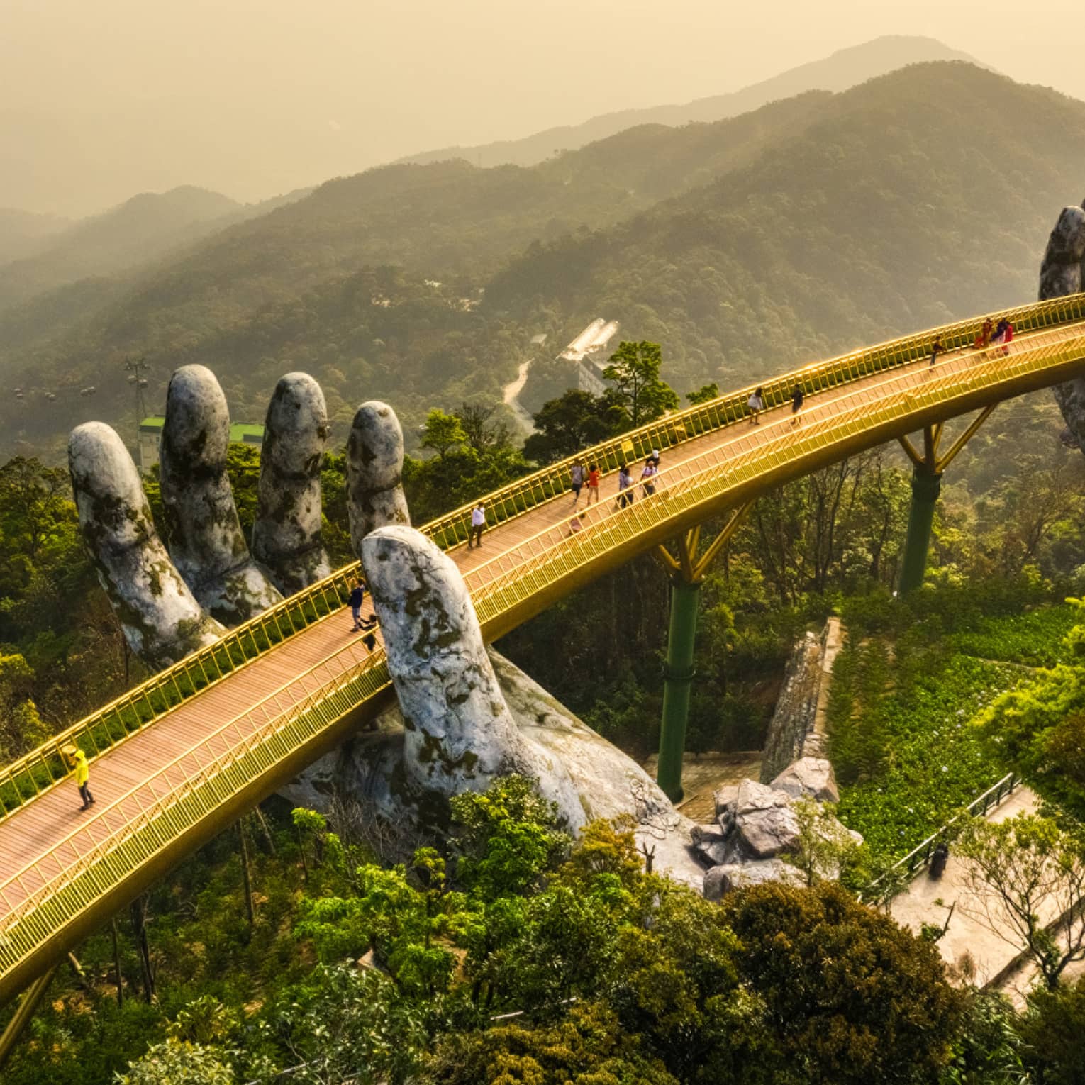 Aerial view of people on a wood-slat bridge above a forest canopy, with golden railings supported by two giant stone hands.