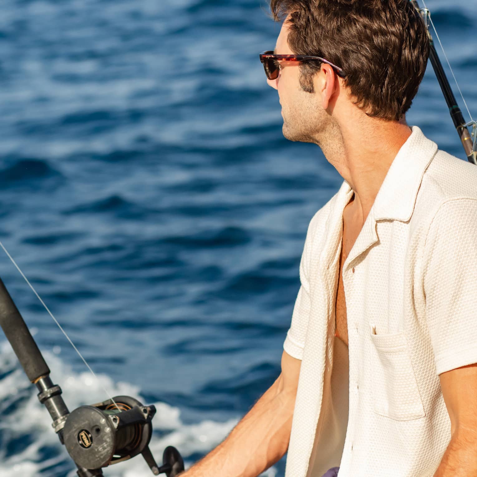 Close-up of a guest wearing sunglasses, sitting beside two offshore fishing rods and looking out towards dark blue waves.
