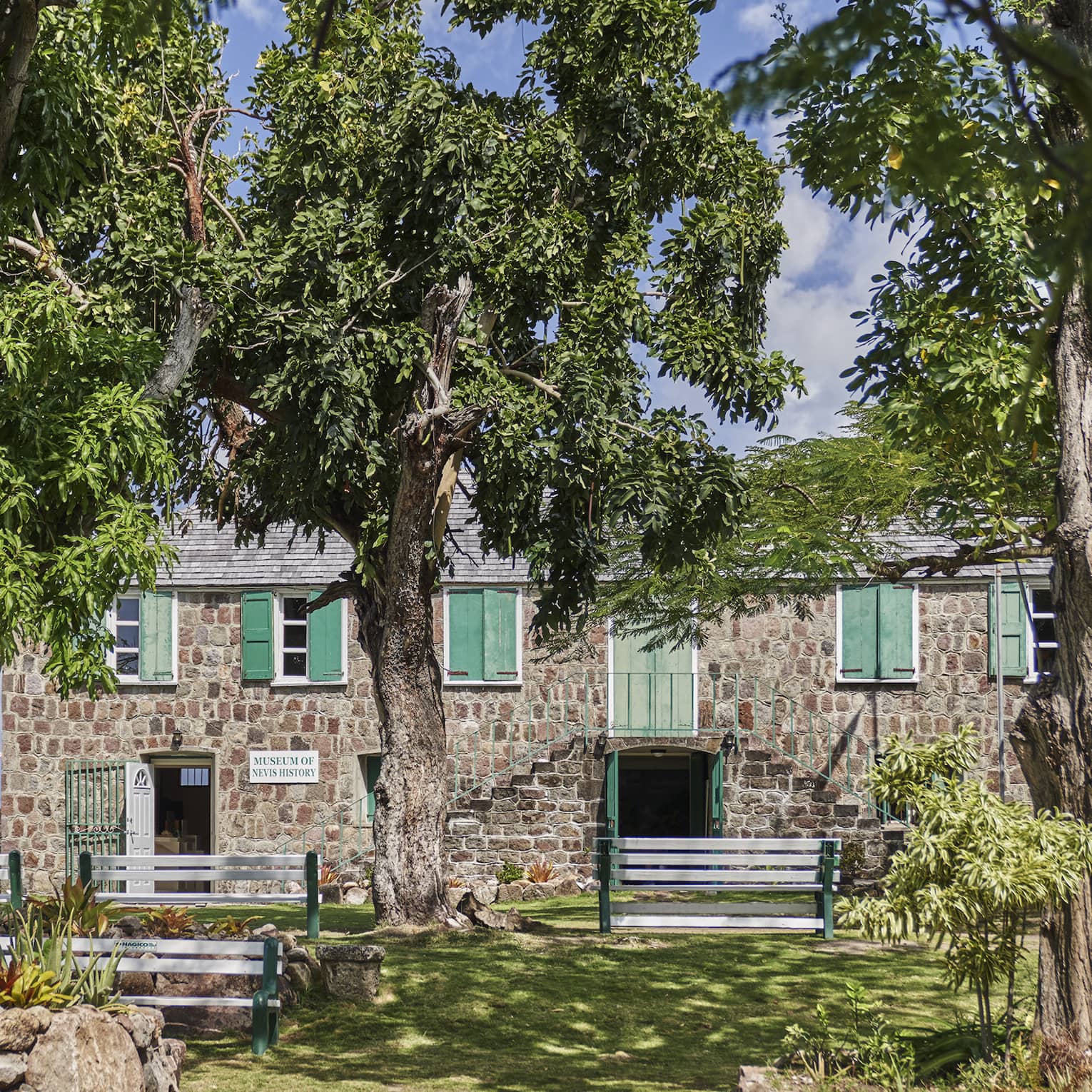 A two-storey stone building with green doors and shutters, seen across a lawn and behind trees, flags blowing behind it.