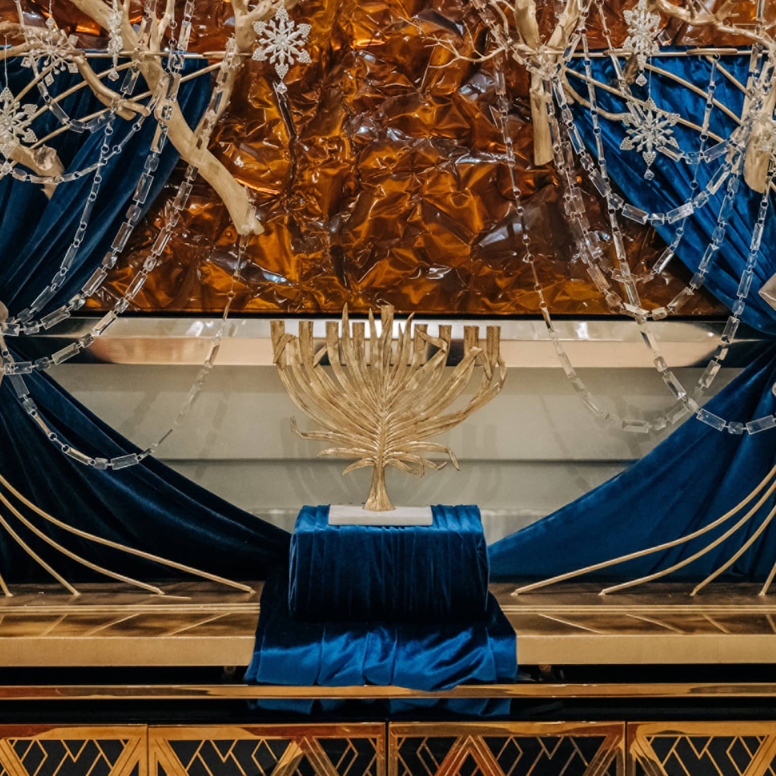 Golden menorah displayed on a blue velvet stand in front of golden tree branches draped in strings of silver