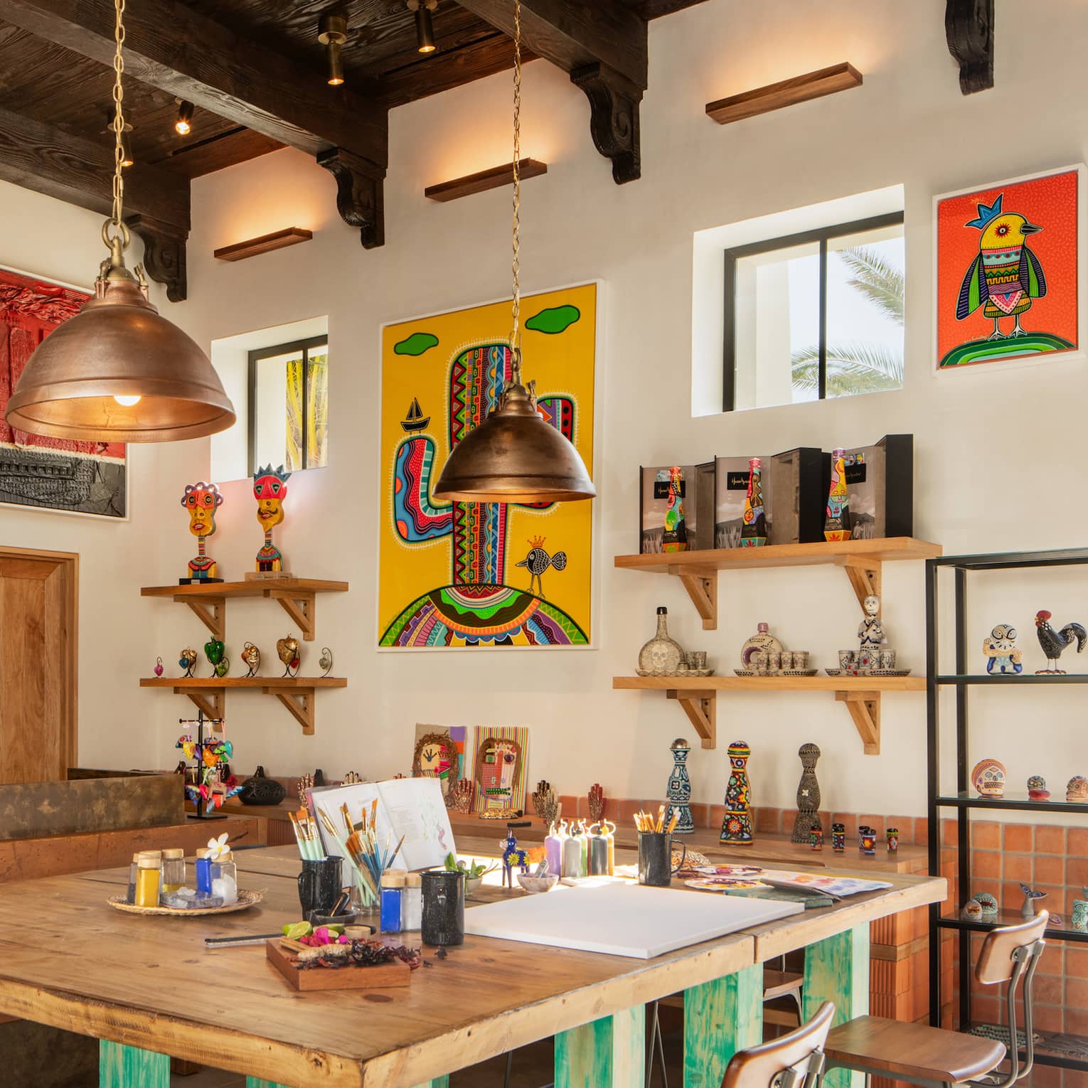 A bright, airy art studio with a large square wooden worktable, shelves of knick-knacks and colourful paintings on the walls.