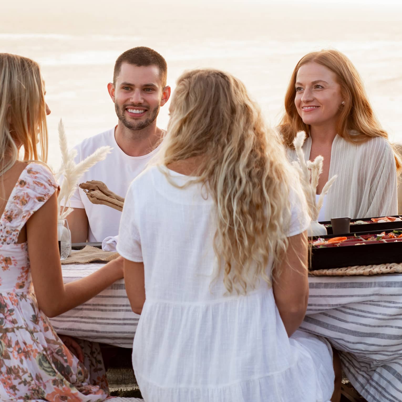 On a cliff overlooking the ocean, a group sits at a picnic table with a striped linen tablecloth and divided picnic boxes.