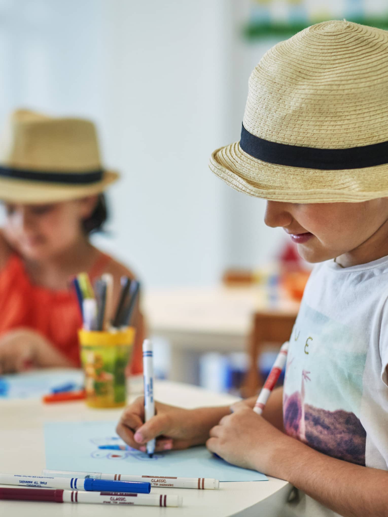 Two smiling children wearing straw fedora hats colour pictures with markers at craft table