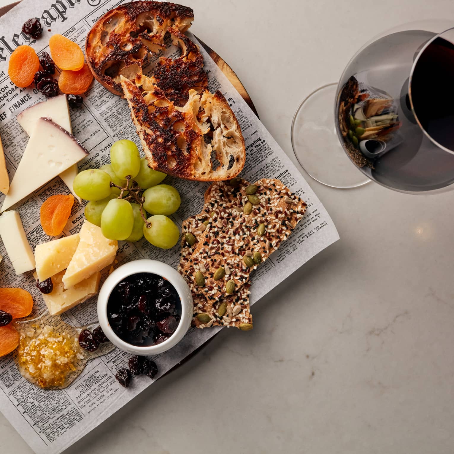 Cheese, grapes, crackers and bread on newspaper on platter next to glass of red wine