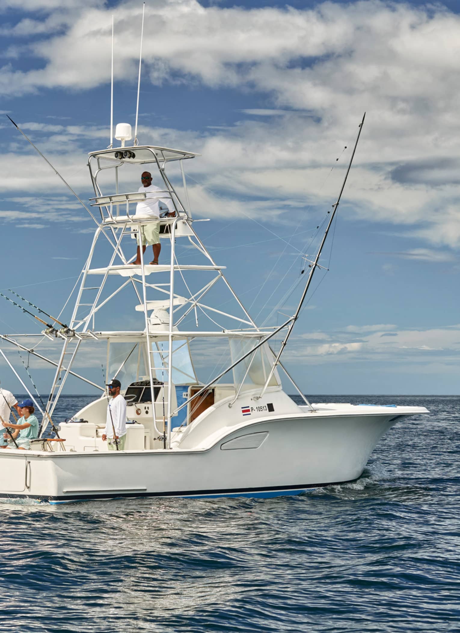 A group of people on a white fishing boat in calm ocean waters, with fishing rods ready. One person is standing on the upper deck, and the sky is partly cloudy.
