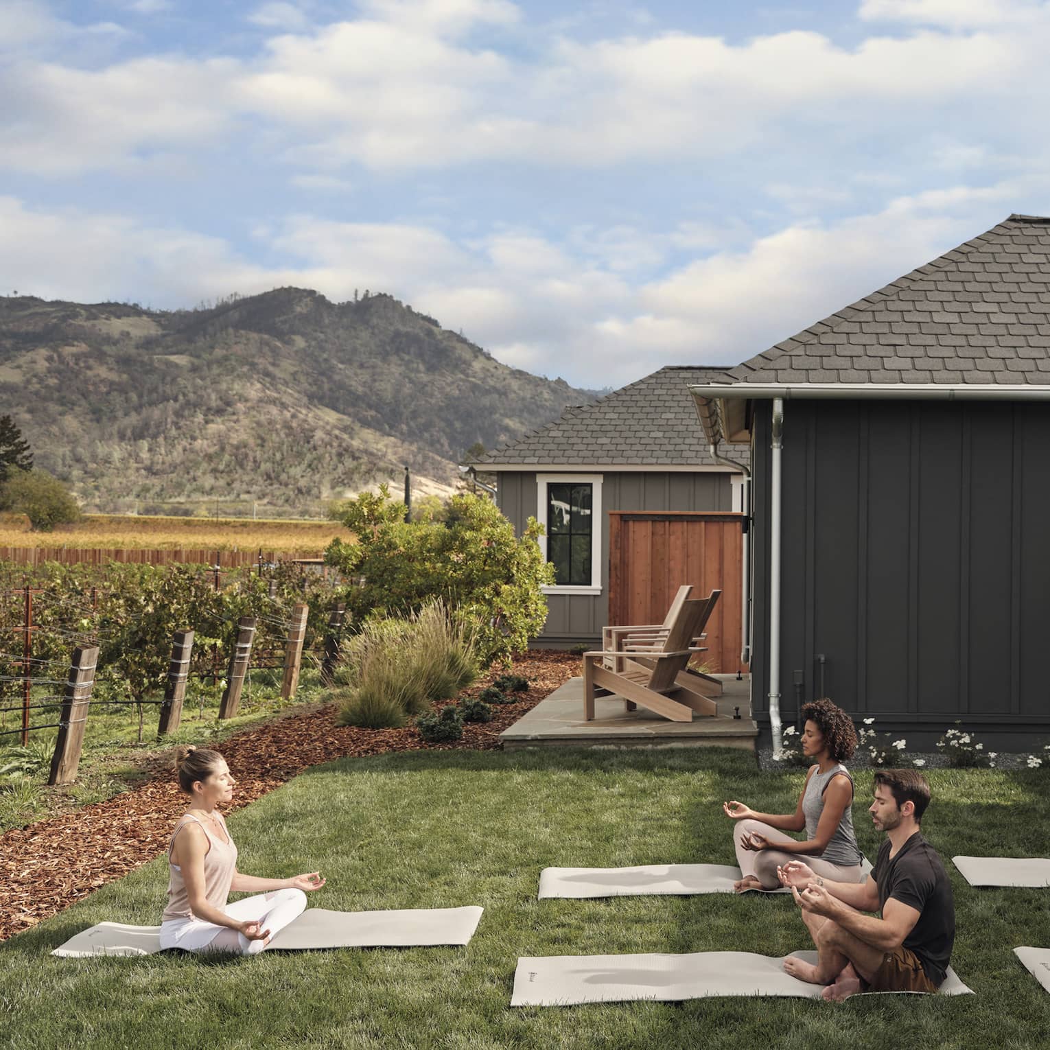 Group of people practicing yoga on mats in a grassy villa backyard near vineyards, with mountain views, wooden chairs and an outdoor grill area under a pergola