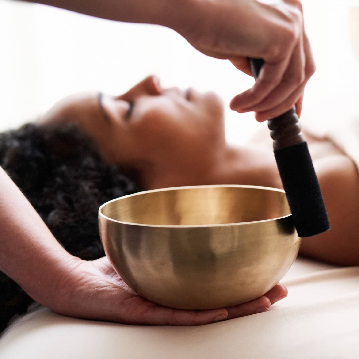 Woman with eyes closed lies on massage table in as vibrational sound therapy is performed with a Tibetan singing bowl