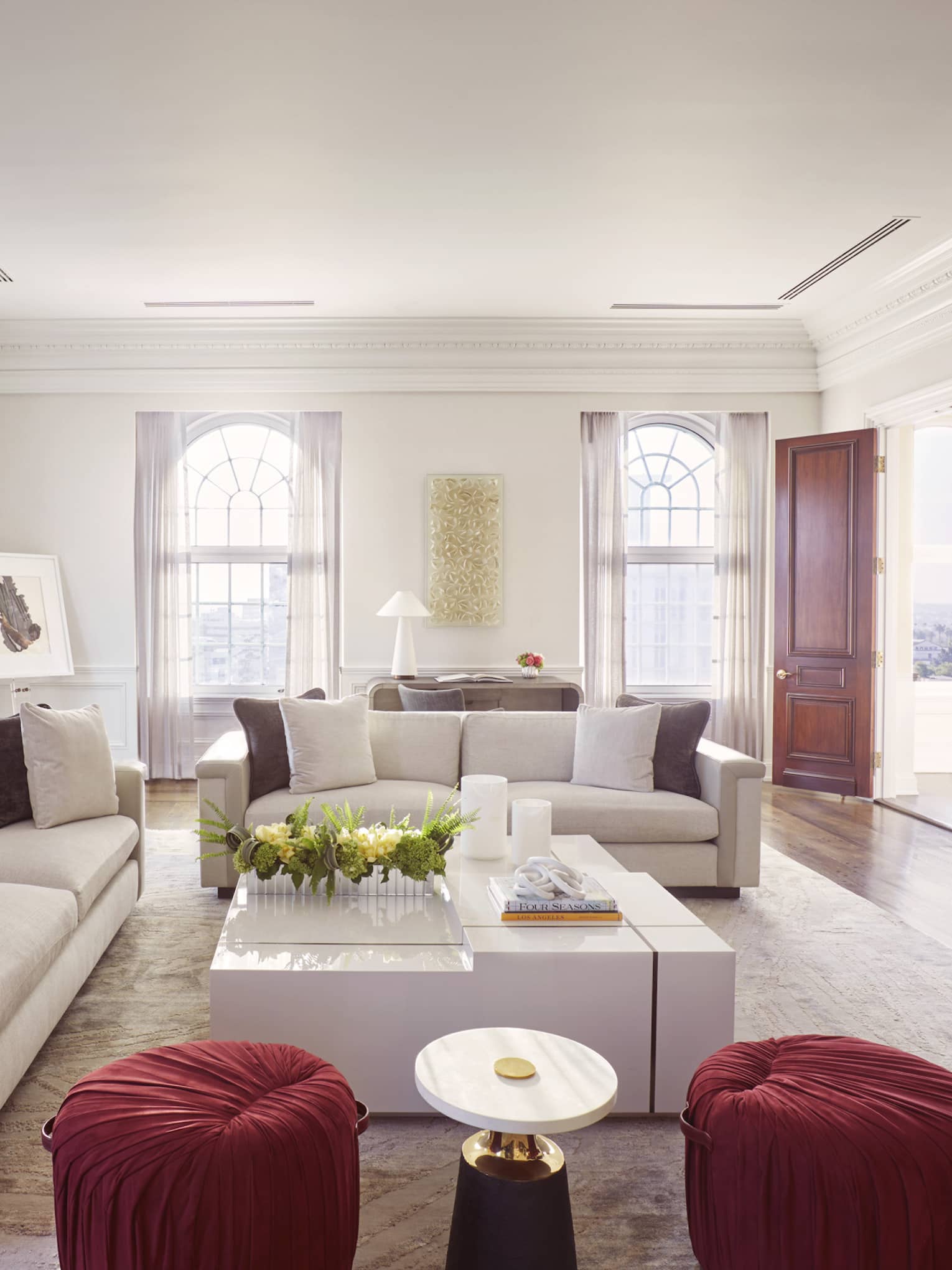Living room with two sofas, large white square coffee table, two red cushioned stools, gold plate artwork on wall, TV and fireplace