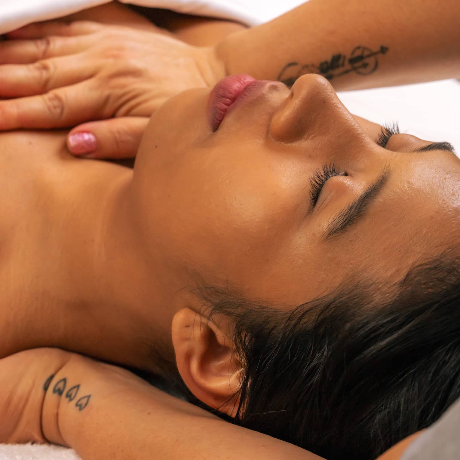 Person lies on their back beneath a white blanket with their eyes closed as a pair of hands massages the back of their shoulder and applies pressure on sternum