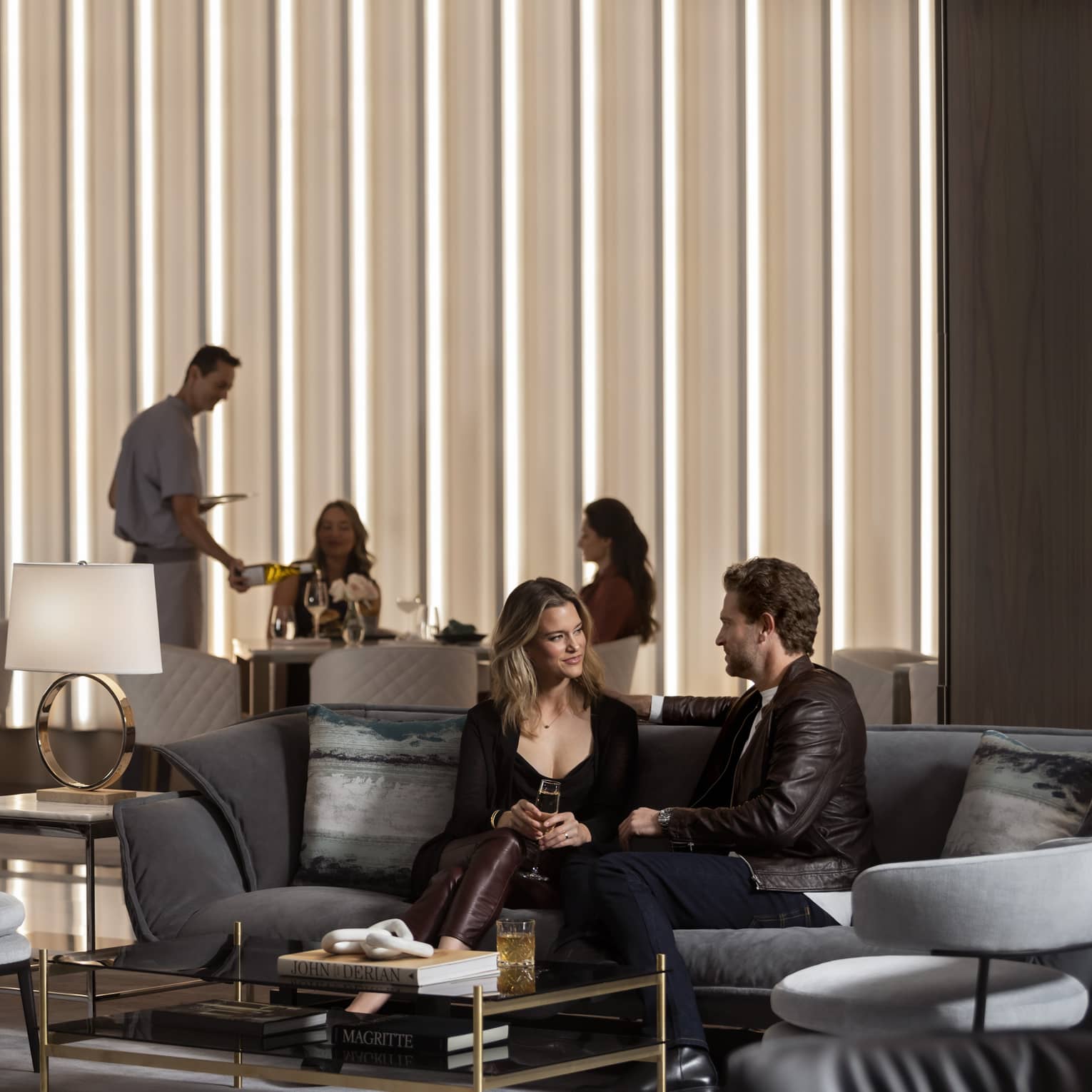 A man and woman sitting on a sofa with a group of people behind them near a large window.