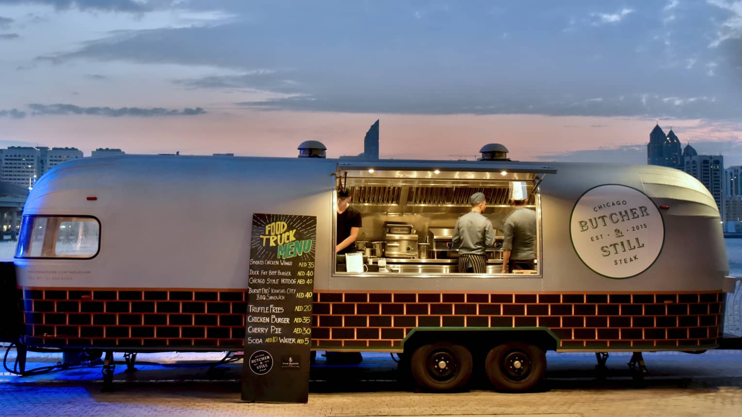 The Butcher and Still airstream food truck is open for business with the door opened, chefs cooking, and sun setting in the distance 