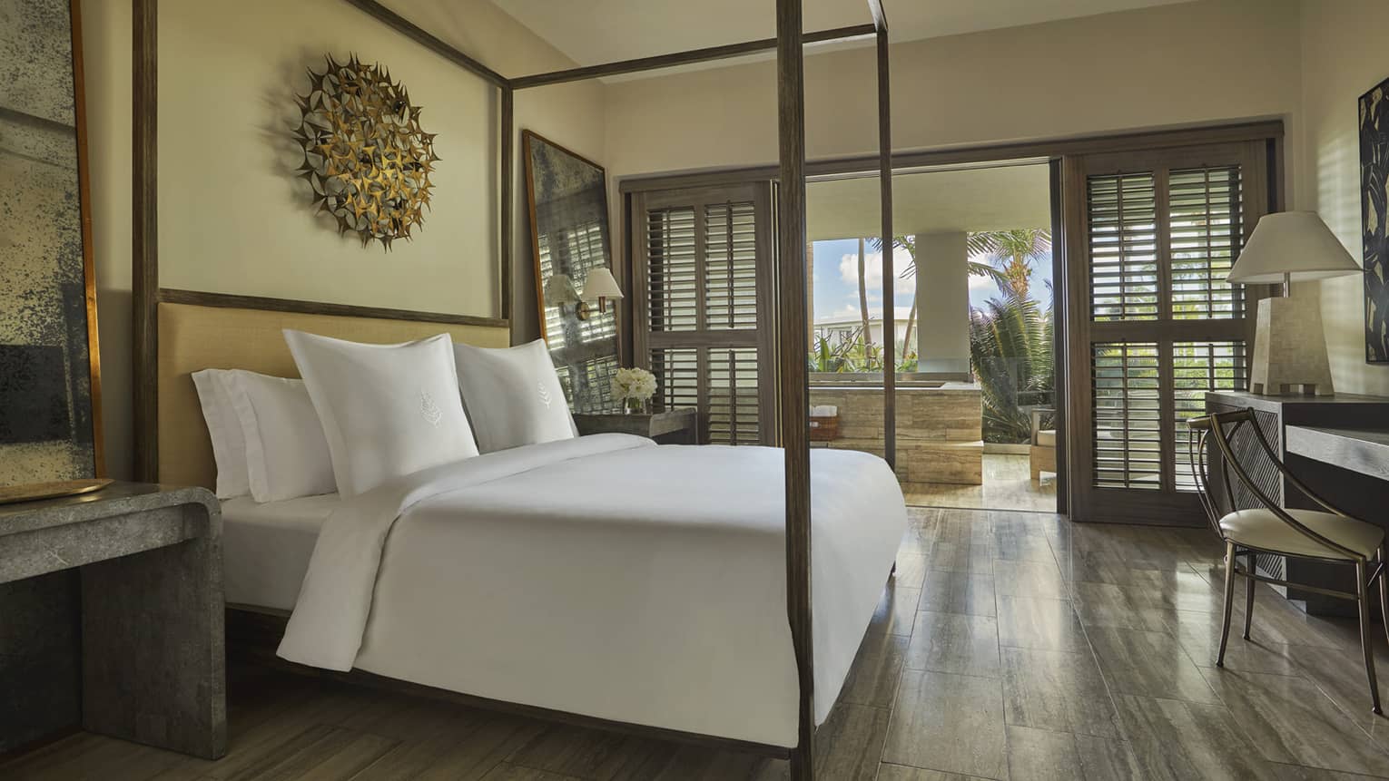 Luxury resort suite bedroom with a four-poster bed, contemporary decor with artwork and a view of a private ocean-view terrace with wooden shutters