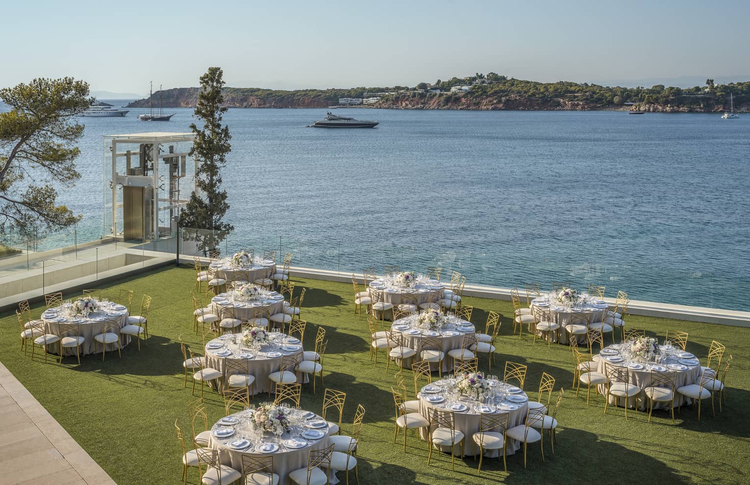 Ten round tables with gold and white chairs and flowers outside on grass overlooking ocean