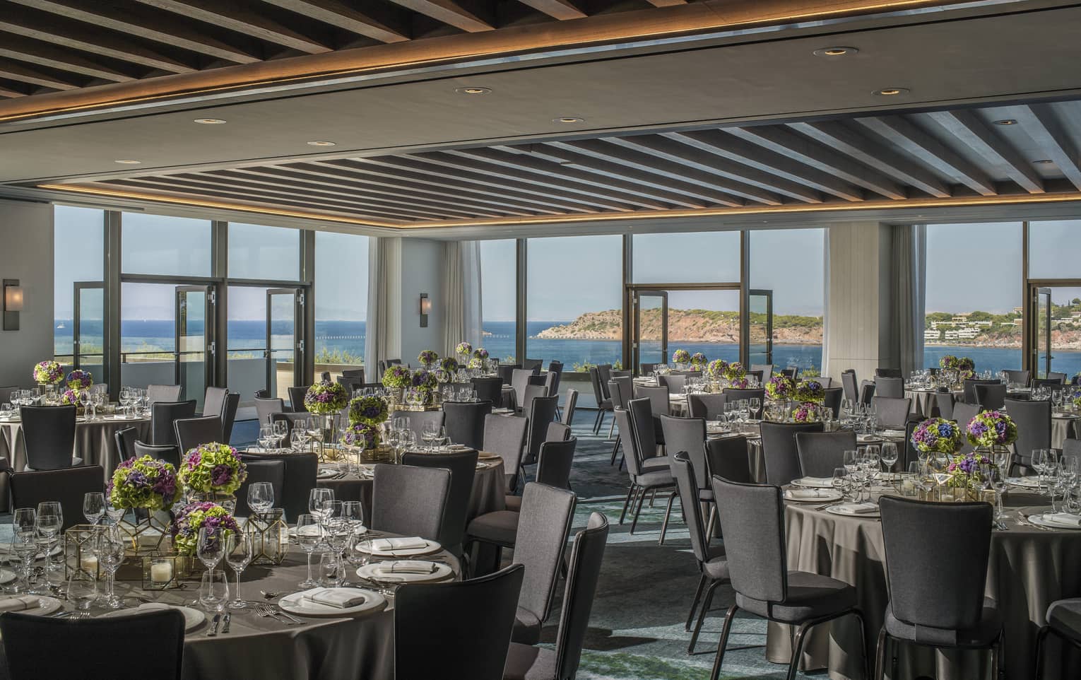 Light filled Nafsika Ballroom with round tables, grey chairs, flowers, wine glasses overlooking ocean 