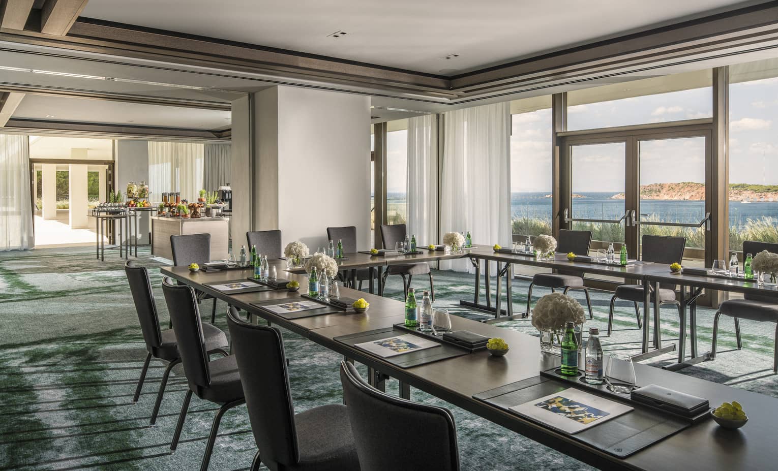 Sunny meeting room with tables in square, water, flowers and buffet, overlooking ocean