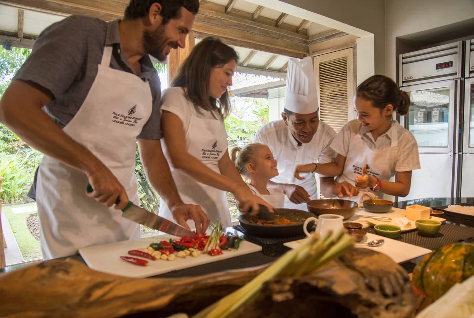 A smiling chef oversees a family joyfully indulging in a private cooking class at Four Seasons