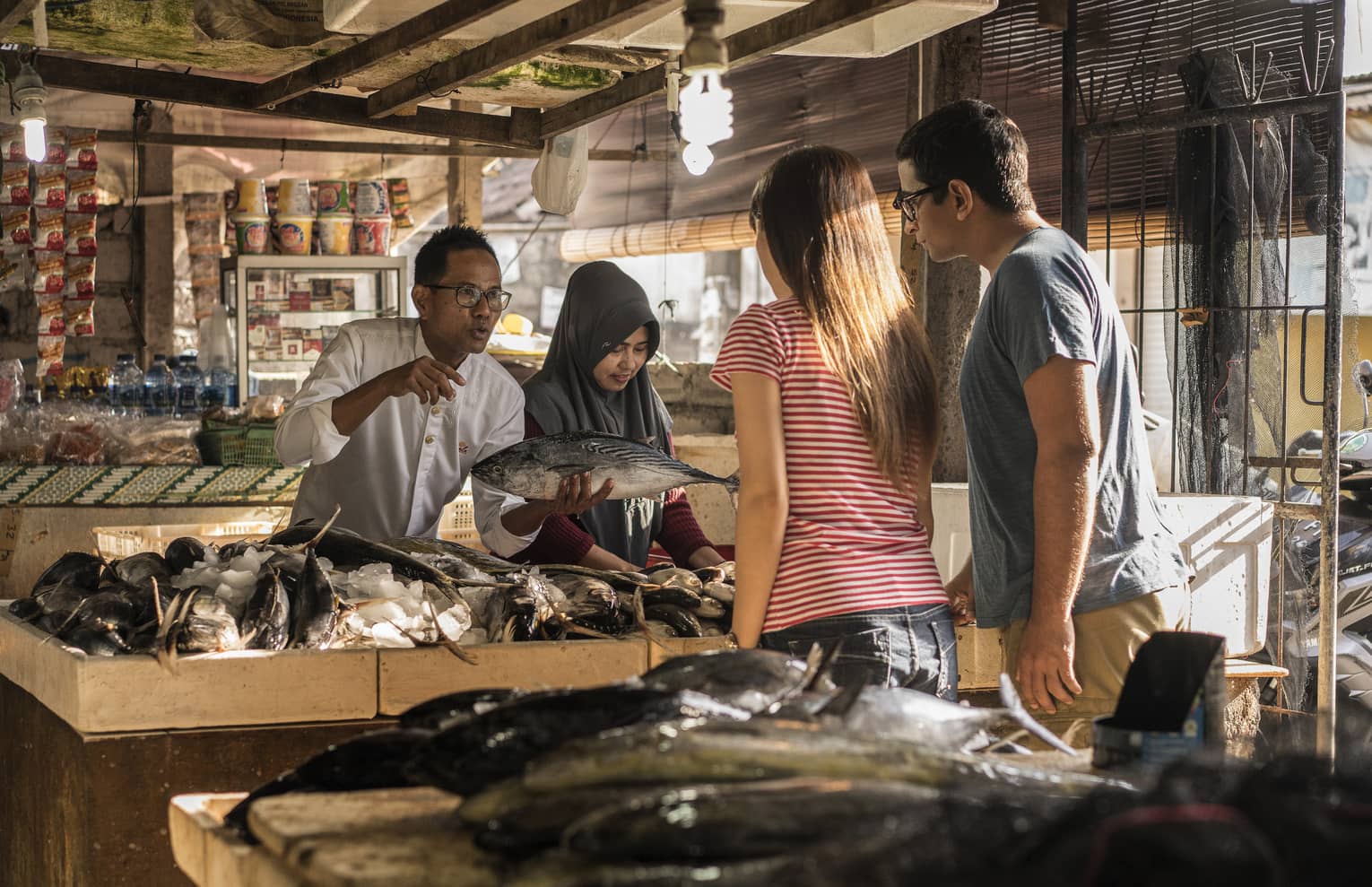 A merchant presents a fish to two tourists before a table of fish in a bed of ice inside a covered market. 